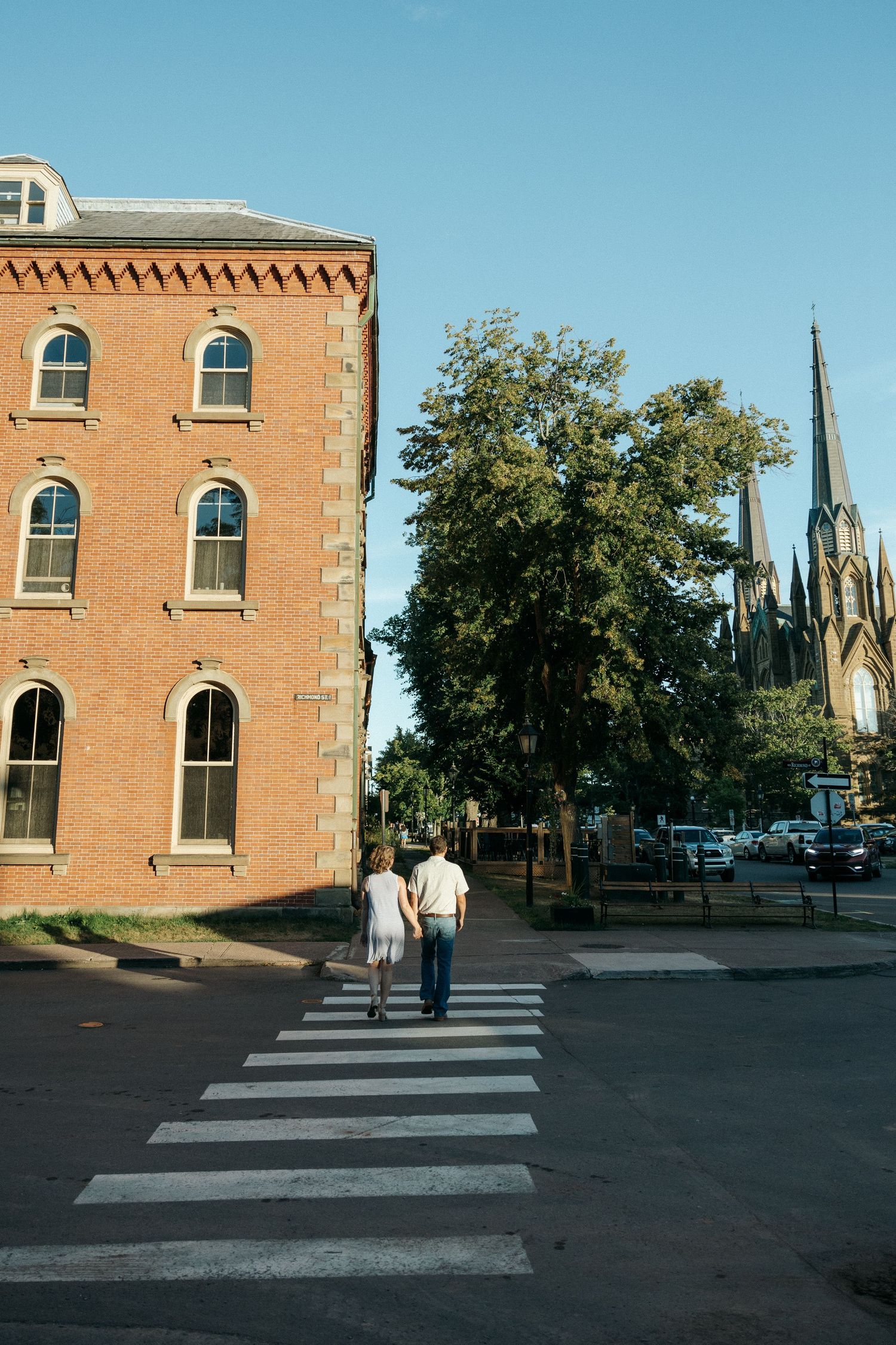 A couple crosses a zebra crossing near a historic brick building on a sunny day.