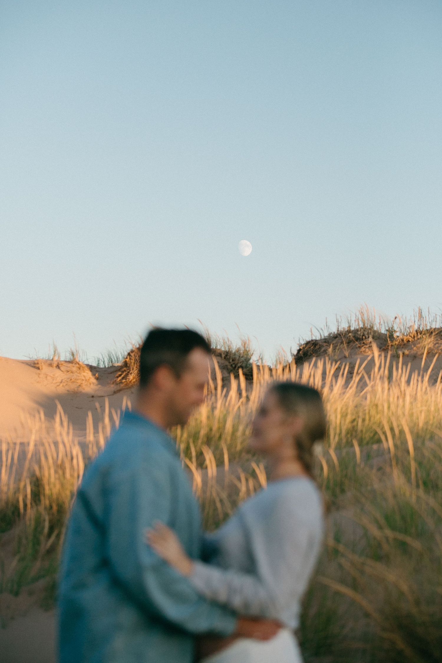 Couple embracing in tall grass with moon visible in blue sky.