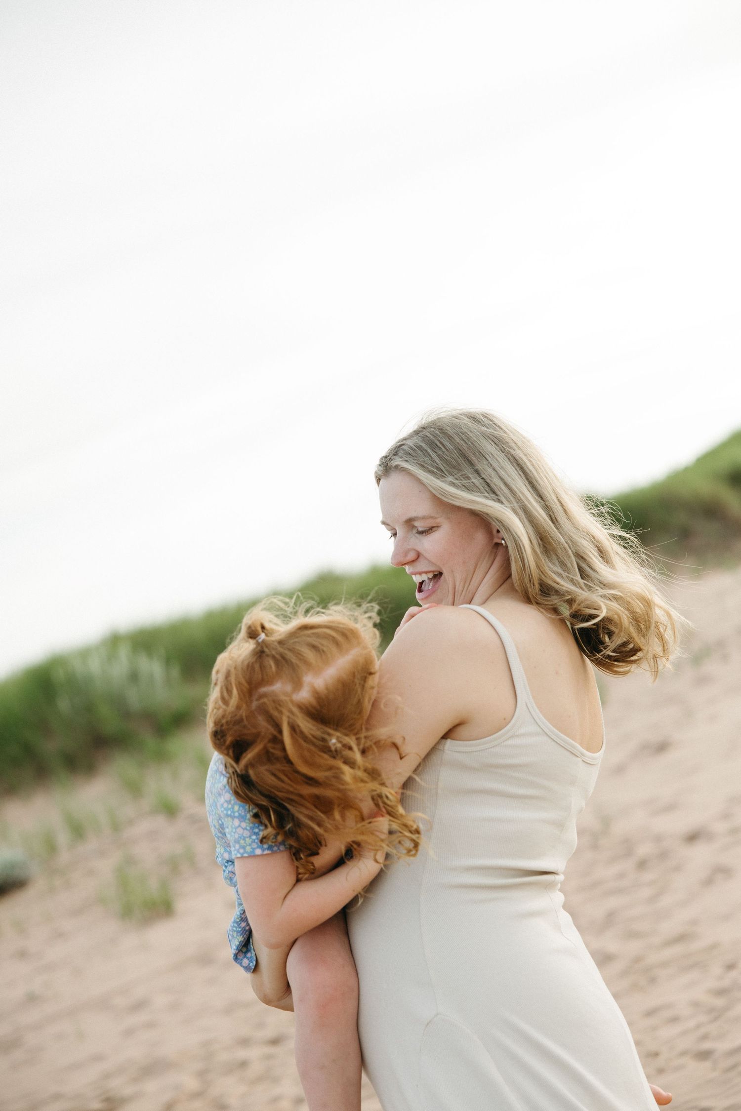 A smiling blonde woman in a white dress hugs a red-haired child on a sandy beach.