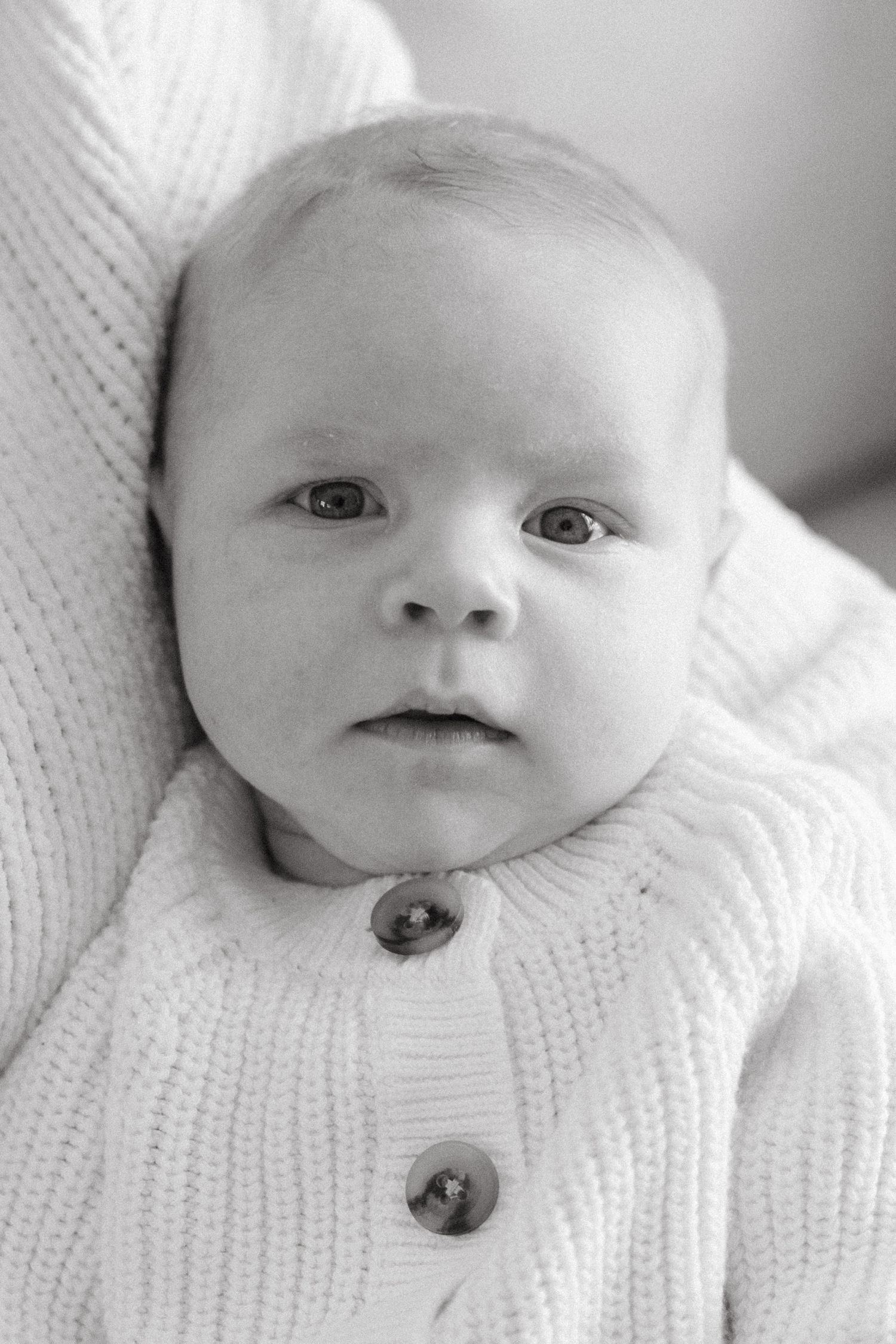 Black and white portrait of a baby wearing a white knit cardigan with buttons while resting on soft fabric.