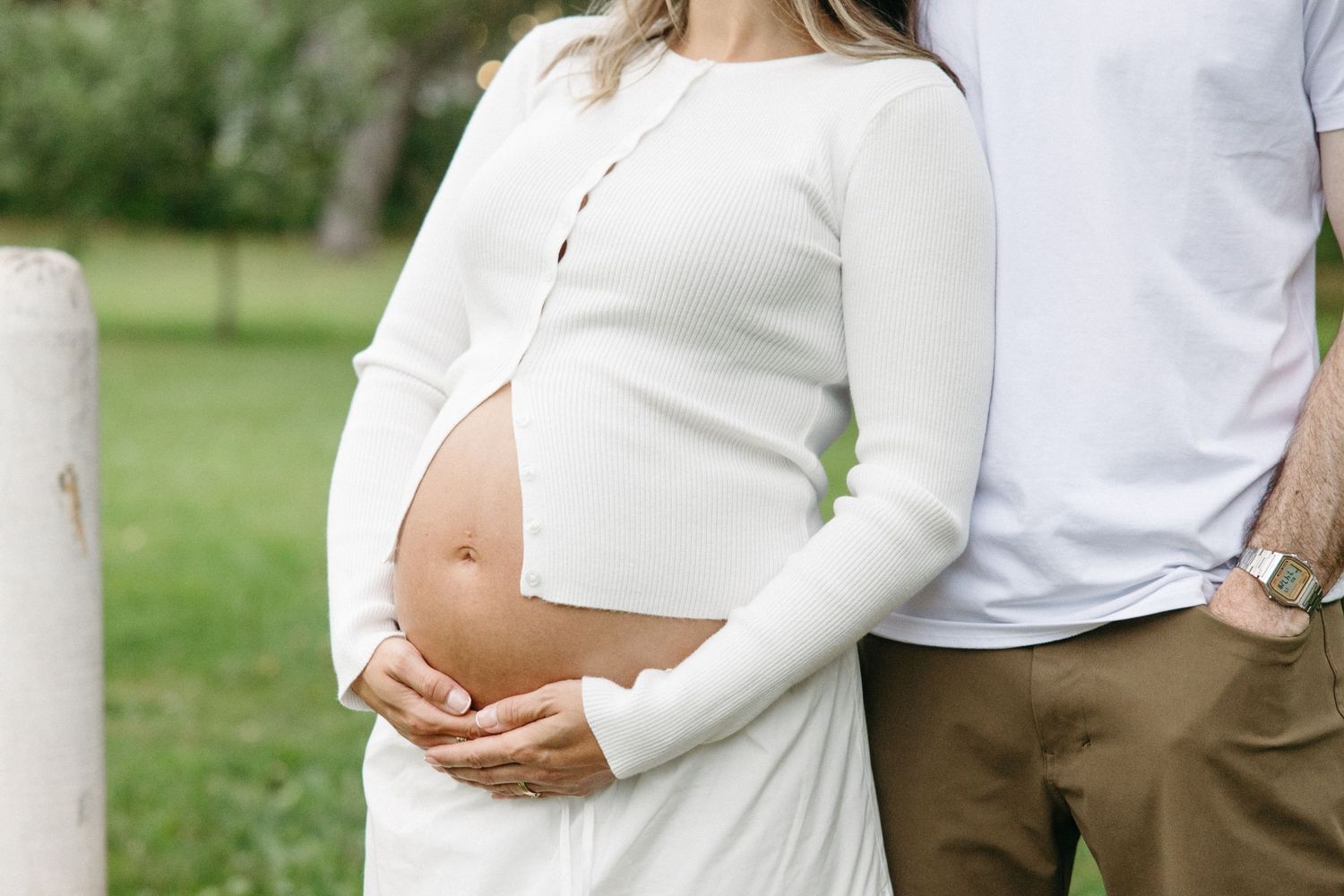 Pregnant woman in white top cradling baby bump outdoors with partner beside her during maternity photo session.