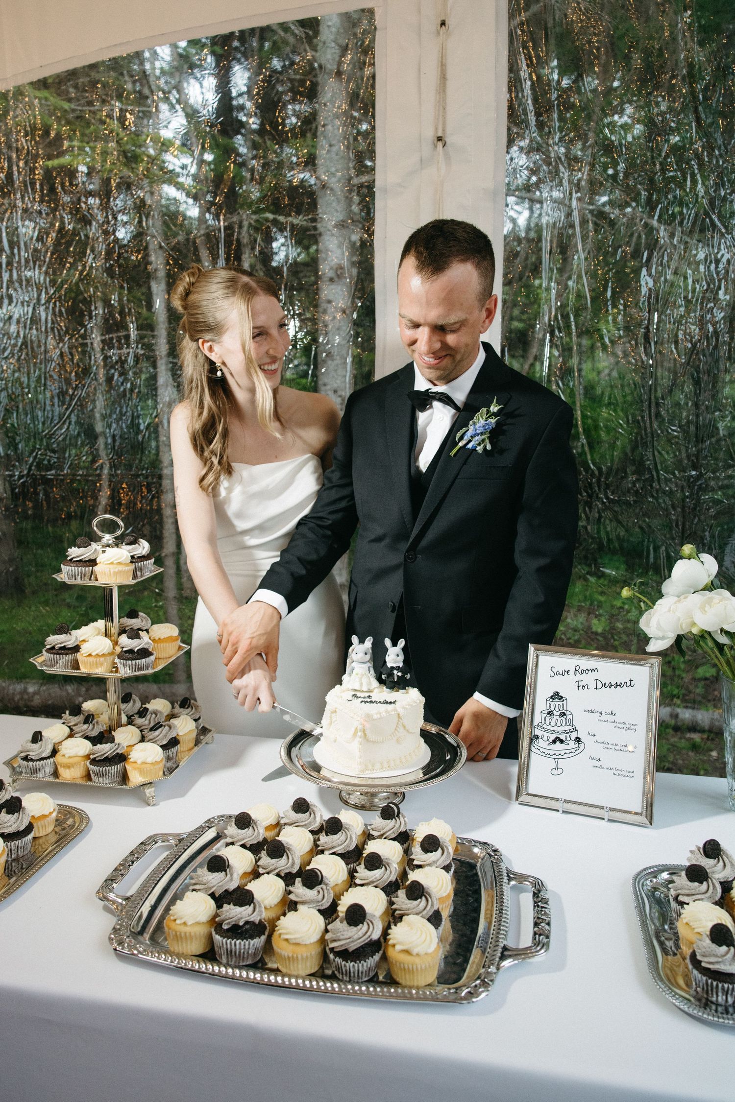 Bride and groom cut small white wedding cake surrounded by cupcakes at reception dessert table.