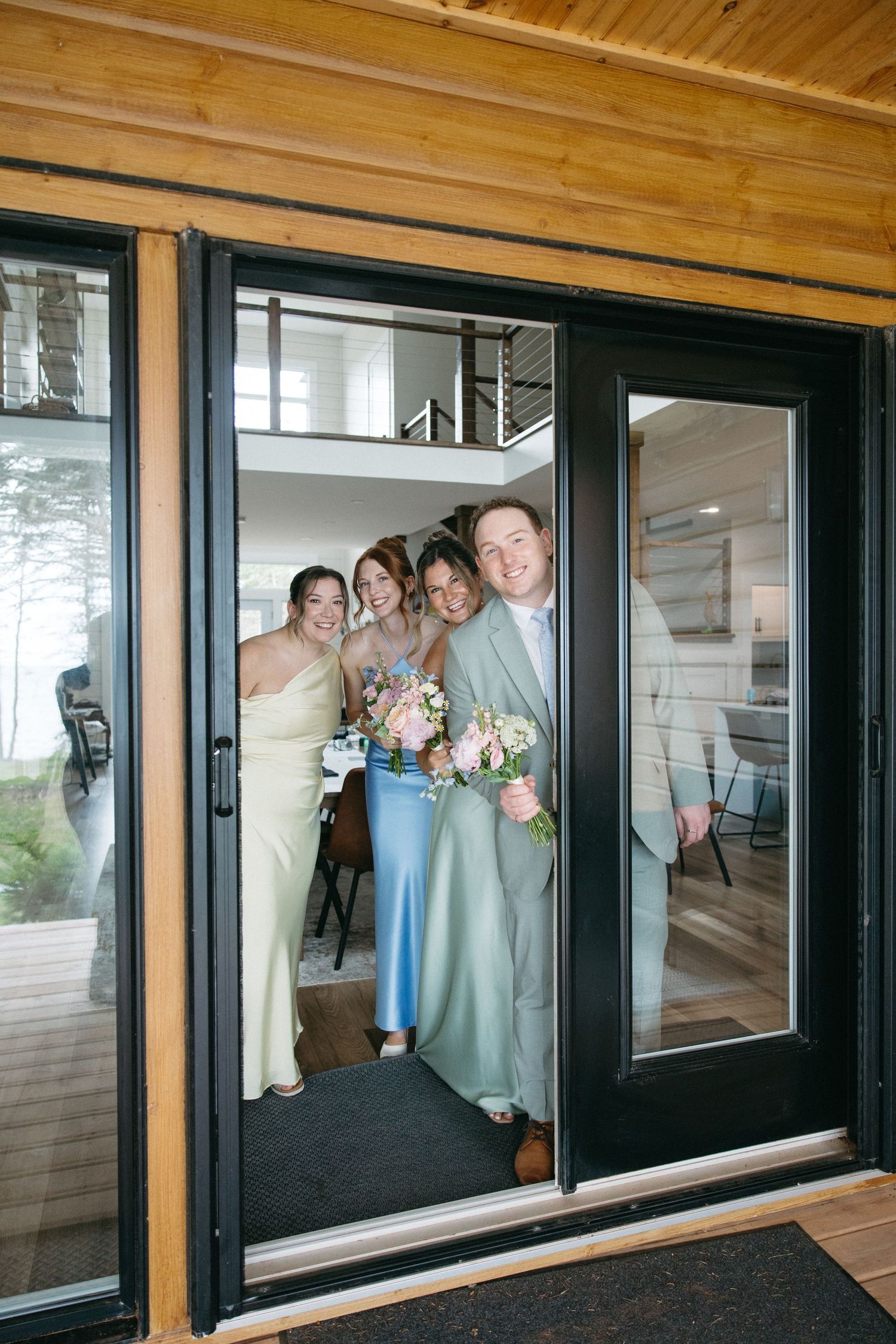 Bridal party smiling and posing in a doorway, bridesmaids in pastel dresses with groom in sage green suit holding bouquet.