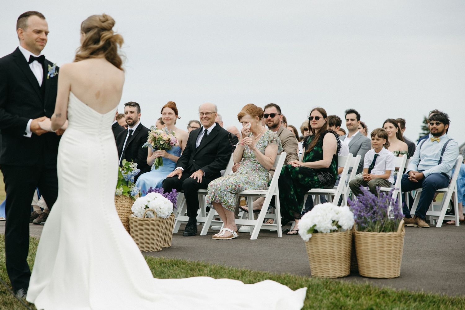 Bride and groom exchange vows at outdoor wedding ceremony while guests watch from white chairs.