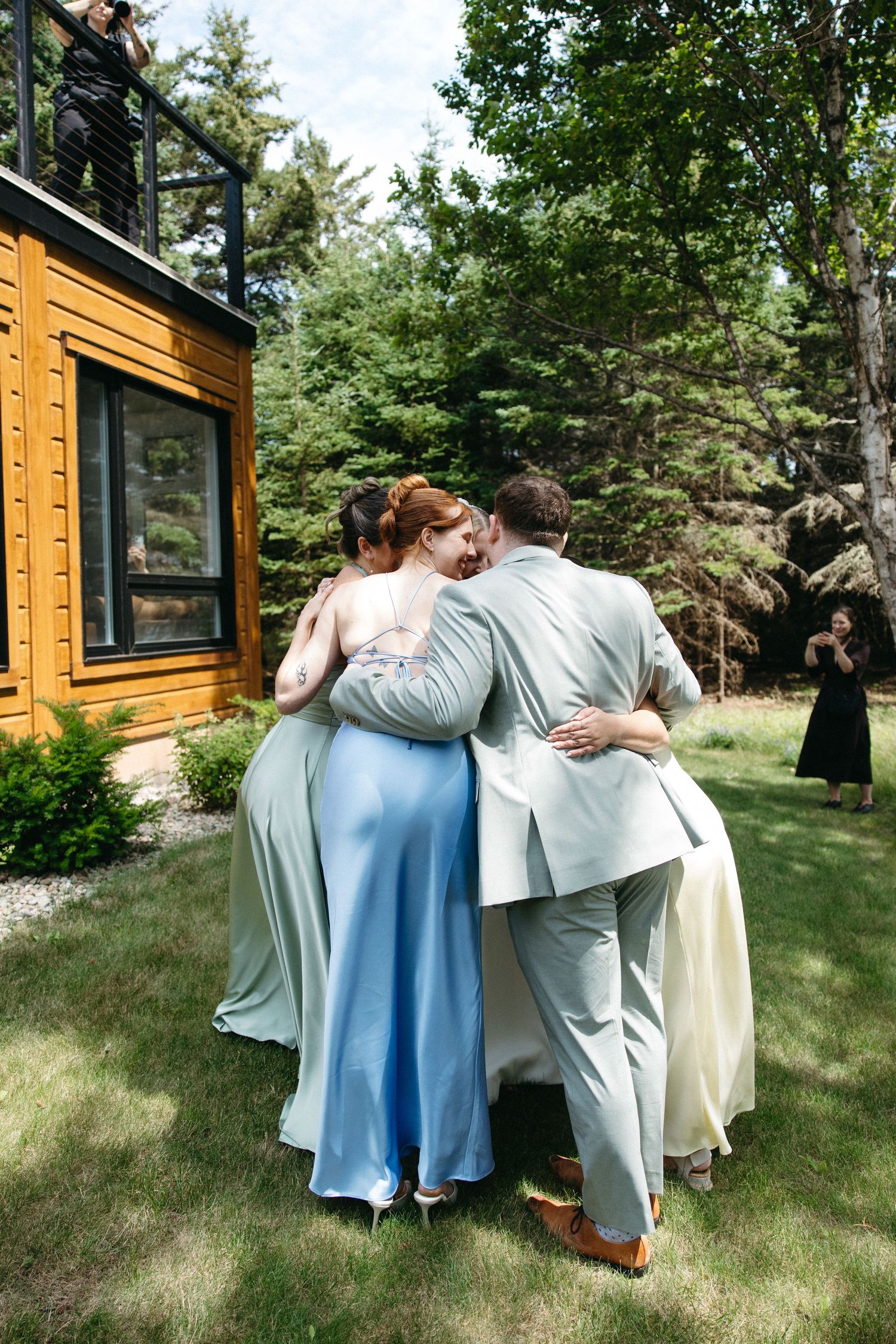 Wedding party group hug outdoors near wooden cabin surrounded by lush green trees on sunny day.