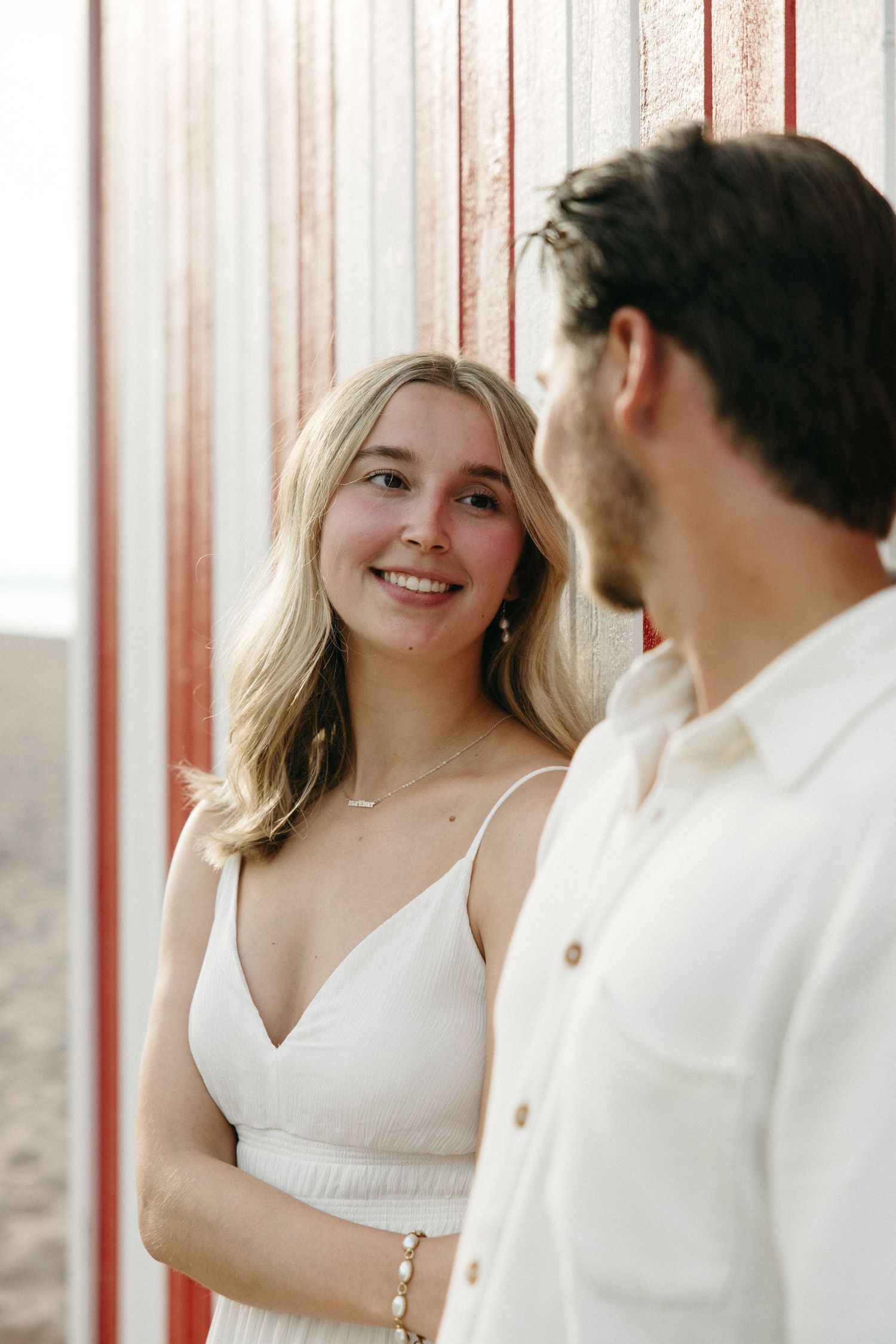Smiling blonde woman in white dress gazing at man near red and white striped wall.