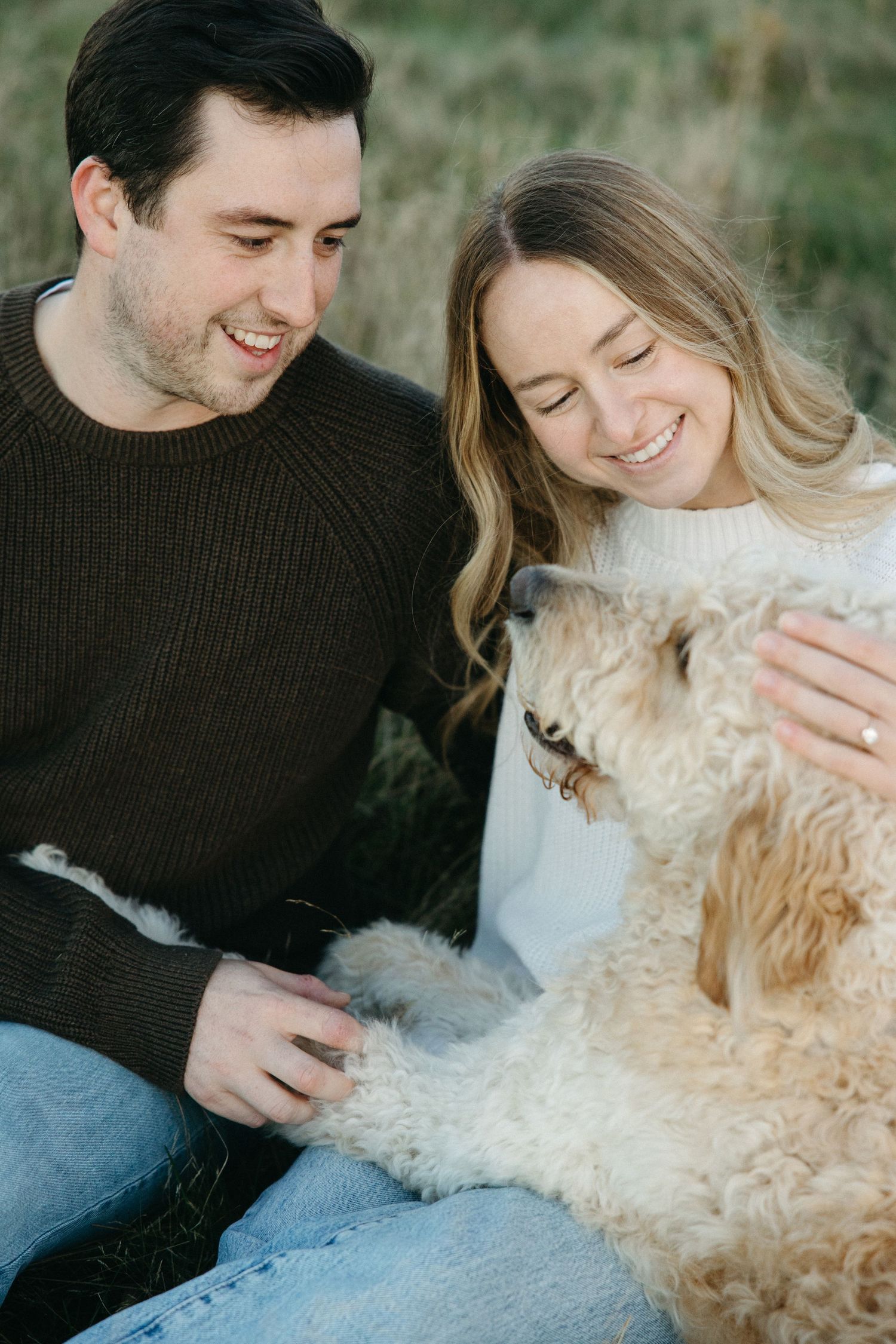 Smiling couple sitting on grass in a field, petting a fluffy golden doodle dog during an outdoor engagement photo session.