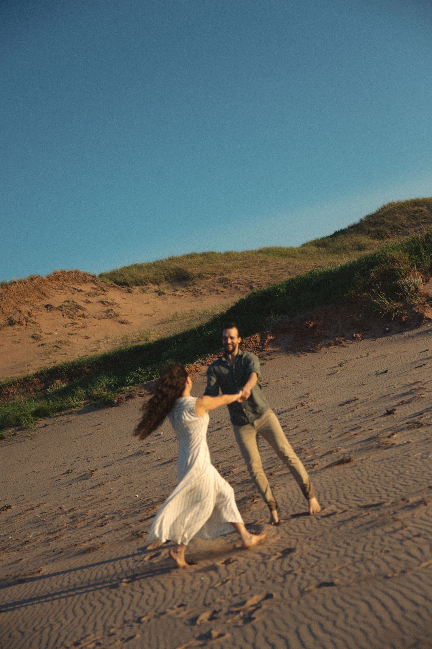 Couple dancing together barefoot on a sandy beach with grass-covered dunes in golden light.