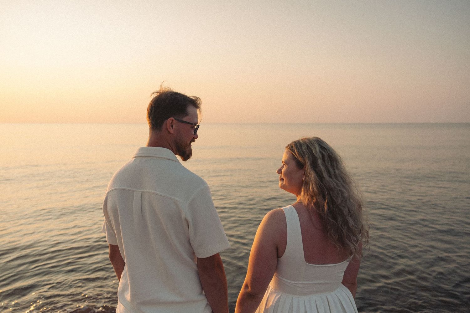 Couple holding hands watching a romantic sunset over the ocean, man in white shirt and woman in white dress.