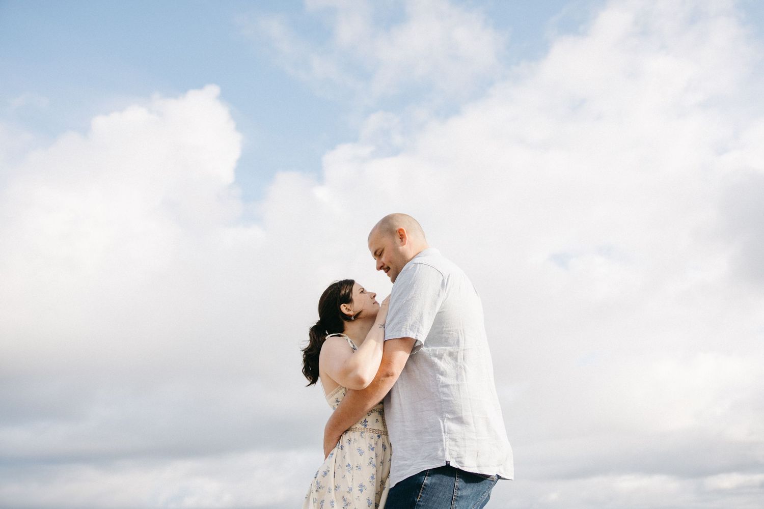 Romantic couple embracing outdoors under a cloudy sky, woman in floral dress and man in white shirt sharing an intimate moment.
