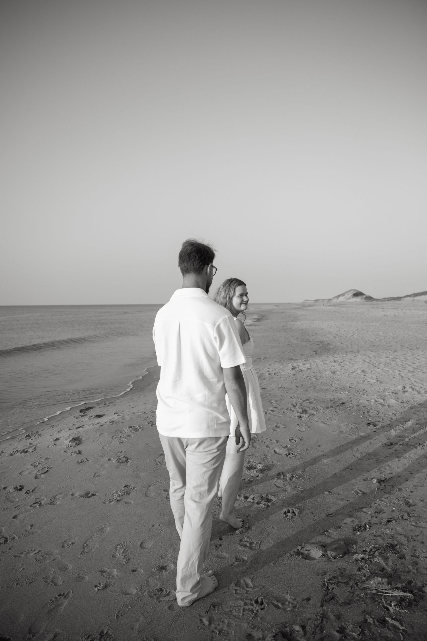Black and white photo of engaged couple walking hand in hand on a sandy beach at sunset.