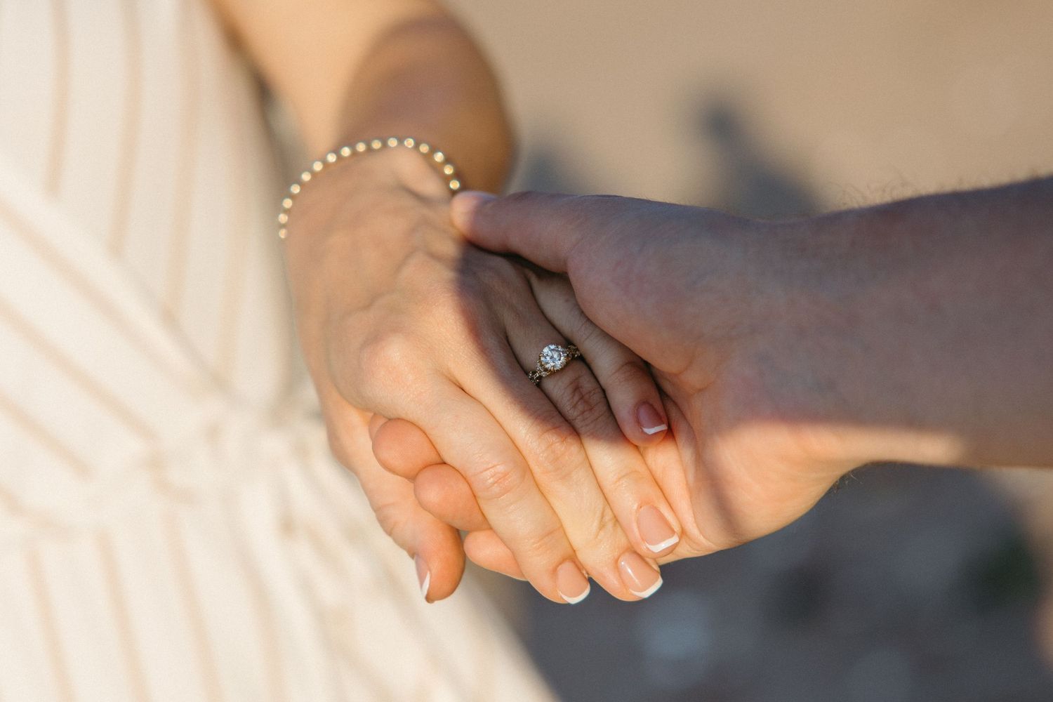 Close-up of an interracial couple holding hands, woman wearing a diamond engagement ring and pearl bracelet.