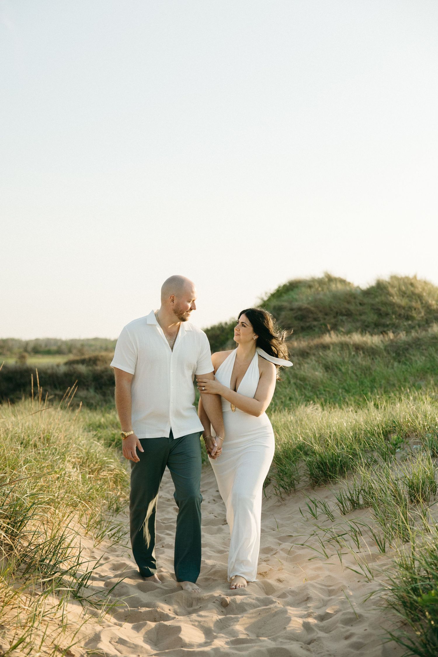 Couple walking hand in hand through sandy beach dunes, woman in white dress, man in white shirt and jeans.