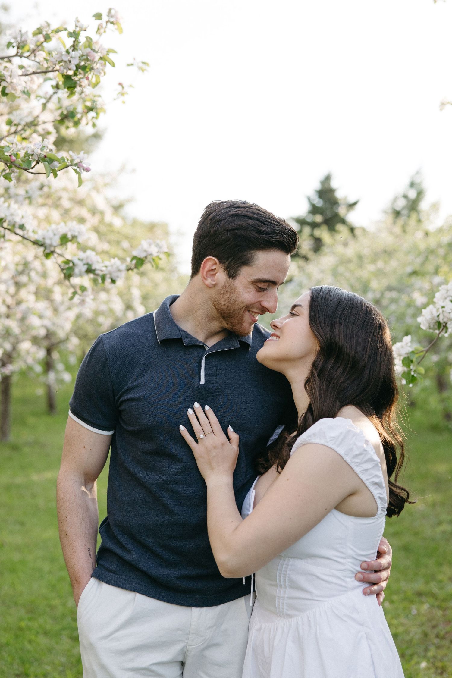 Couple sharing a tender almost-kiss moment surrounded by white blossoming trees in spring orchard.