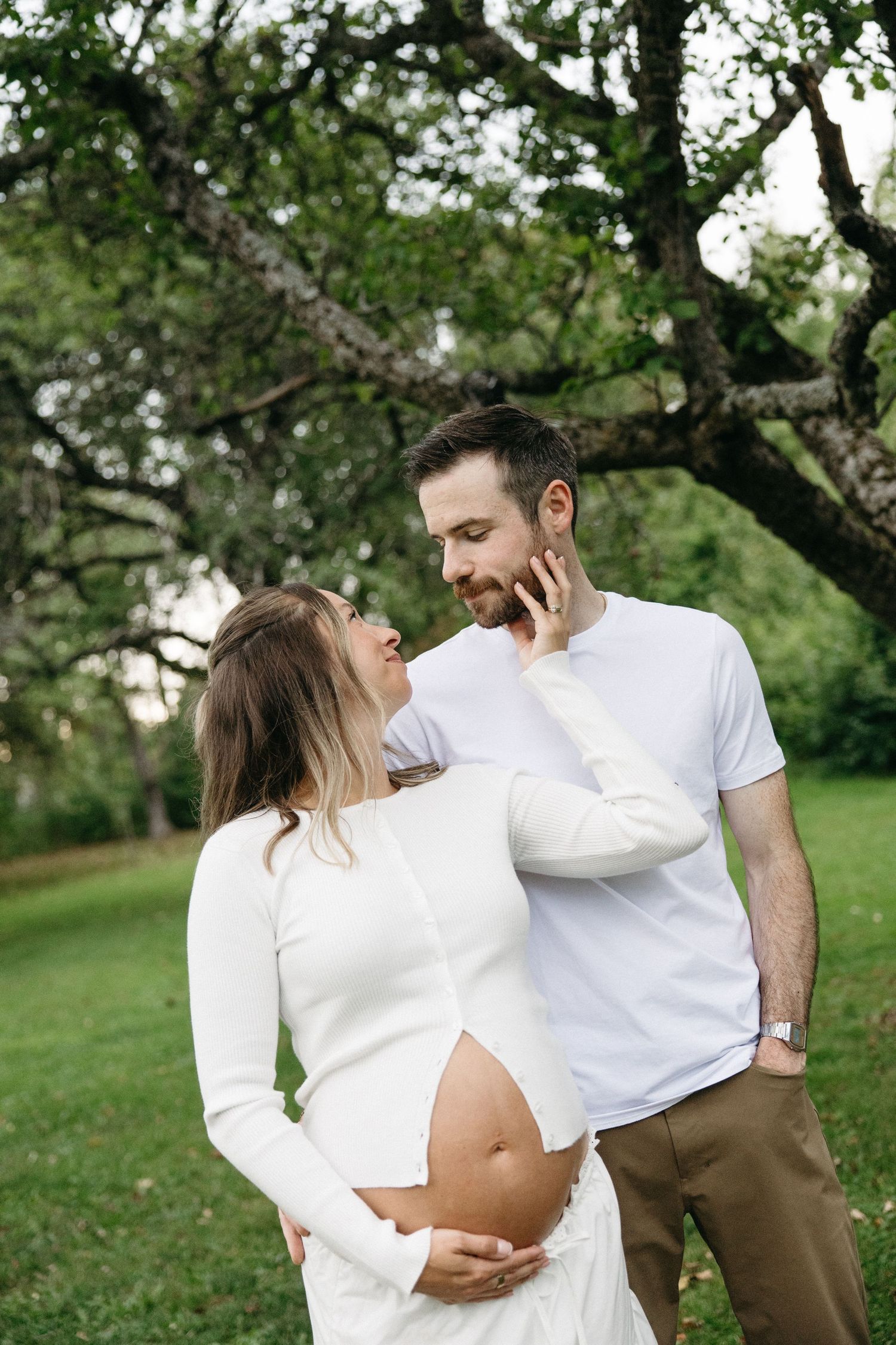 Pregnant couple in white outfits share a tender moment outdoors, woman touching mans face while cradling her baby bump.