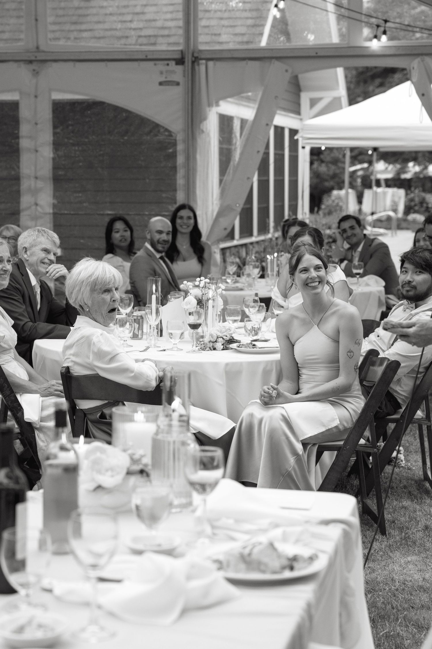 Black and white photo of wedding guests laughing and enjoying speeches during an elegant tented reception.