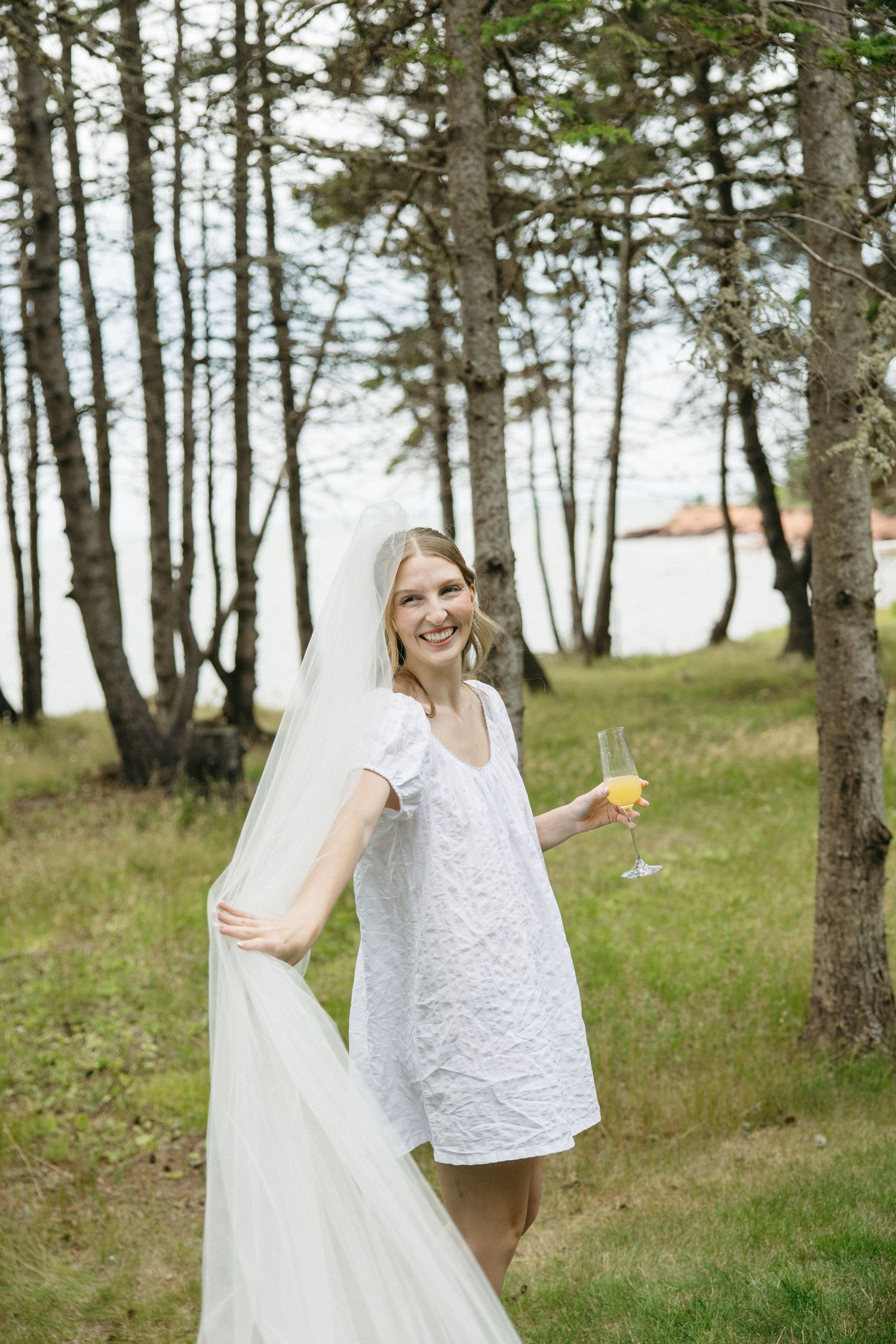 Smiling bride in white mini dress and veil holds champagne glass outdoors among tall trees near water.