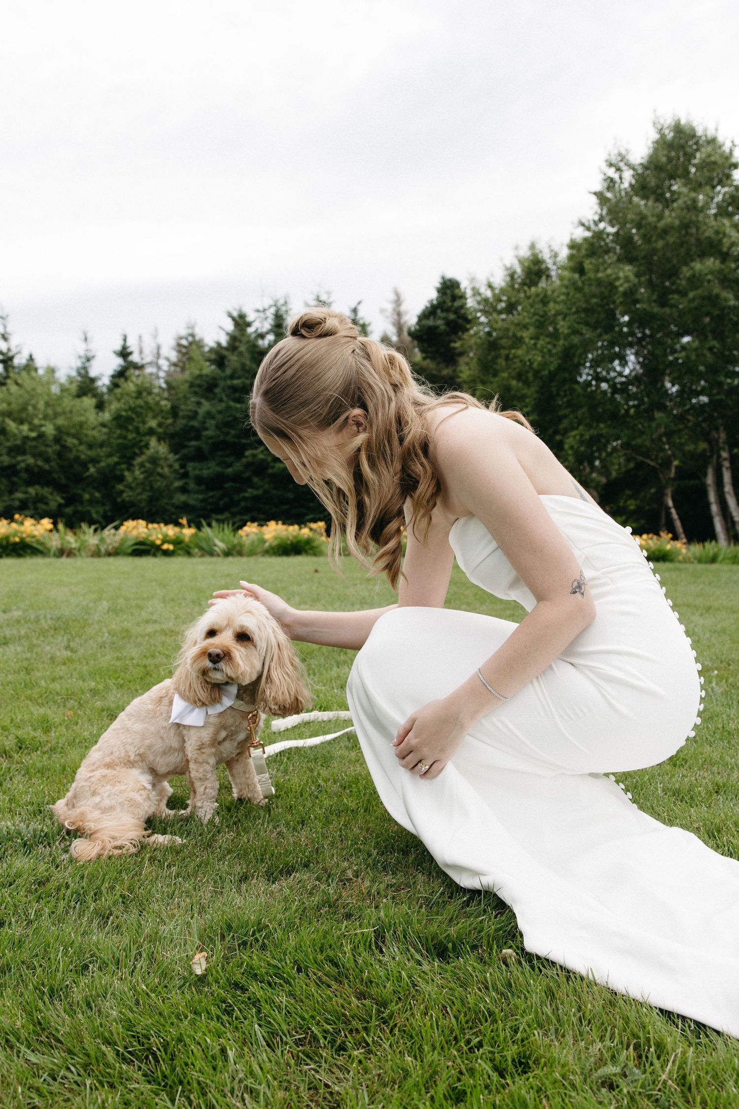 Bride in white gown crouches on grass to pet small dog wearing bow tie at outdoor wedding.