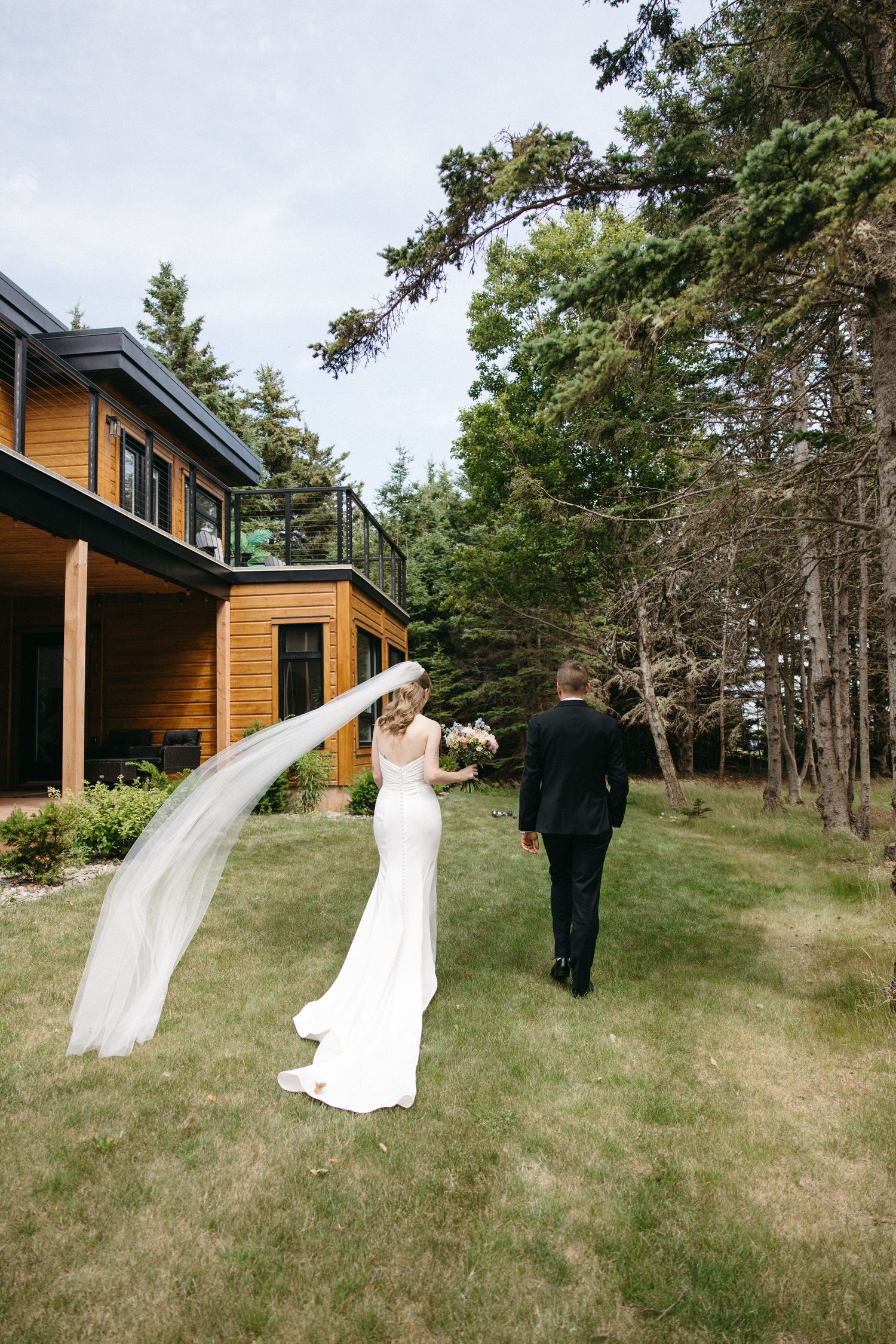 Bride and groom walk hand in hand across lawn with veil flowing near wooden venue.