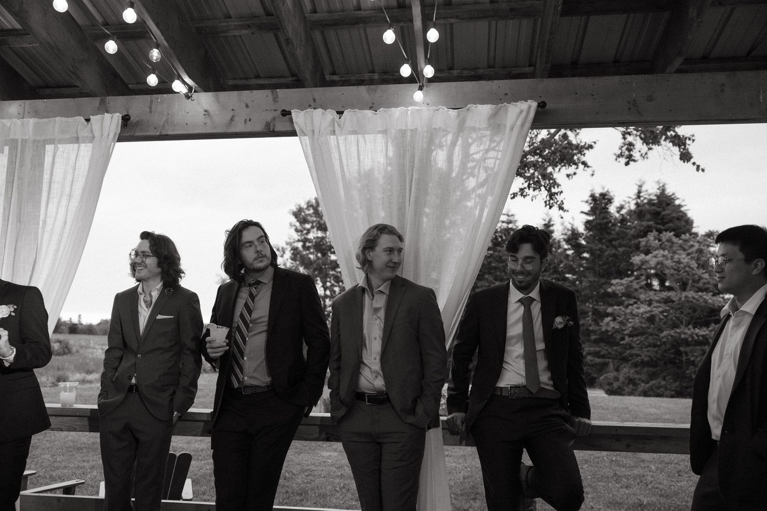 A group of groomsmen pose casually under white drapes at an outdoor wedding venue.