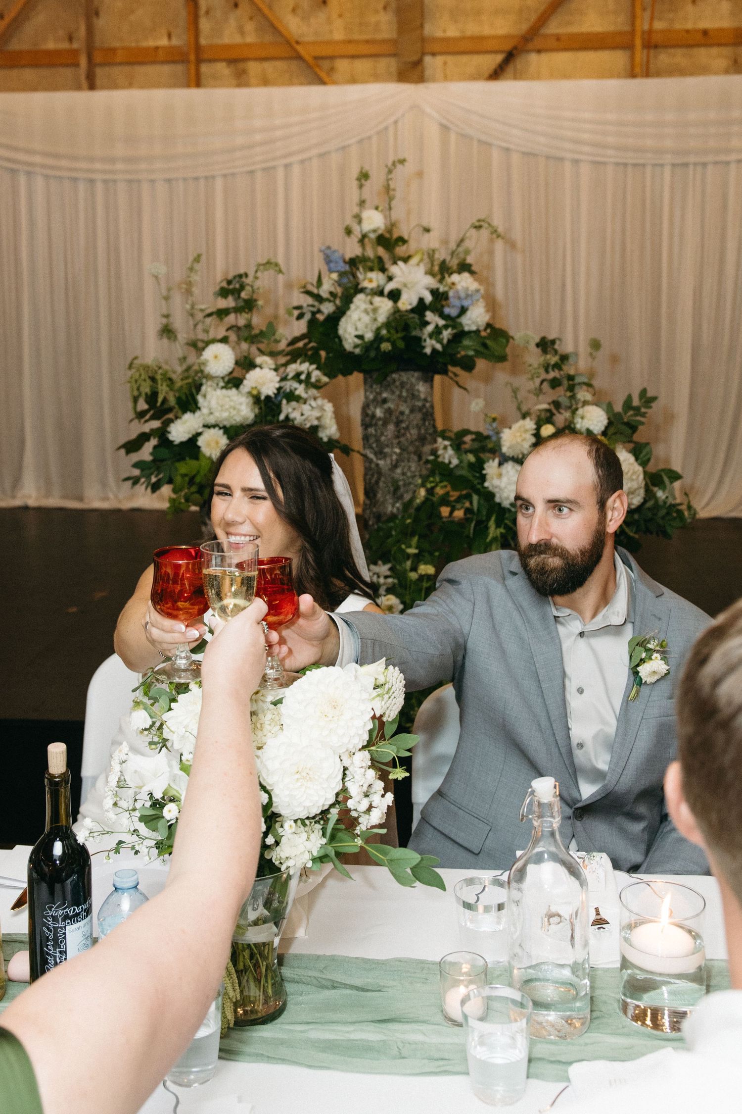 Wedding party members seated at decorated tables with white floral centerpieces in a rustic venue.
