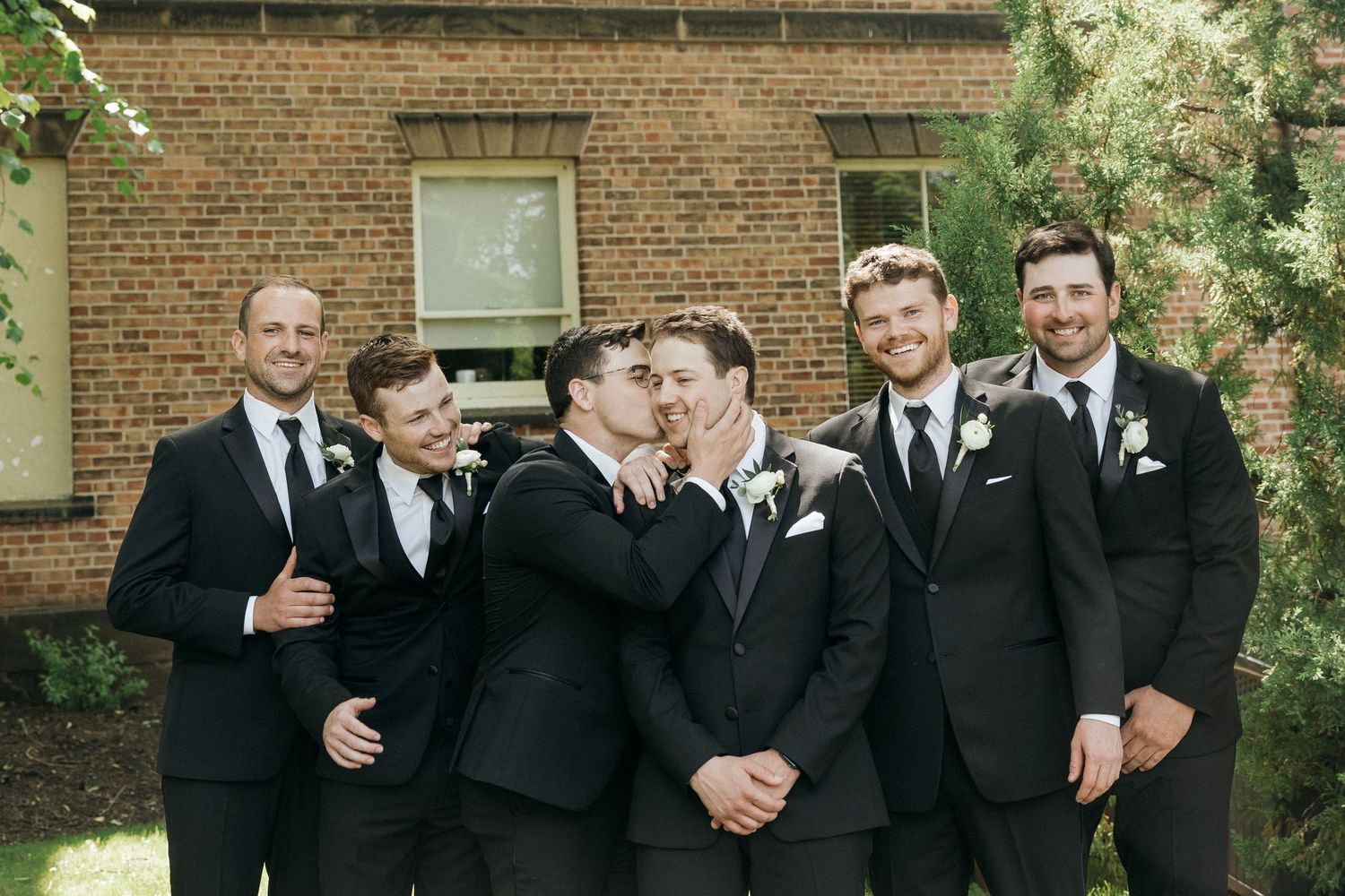 Groomsmen in black suits pose together against brick wall backdrop.