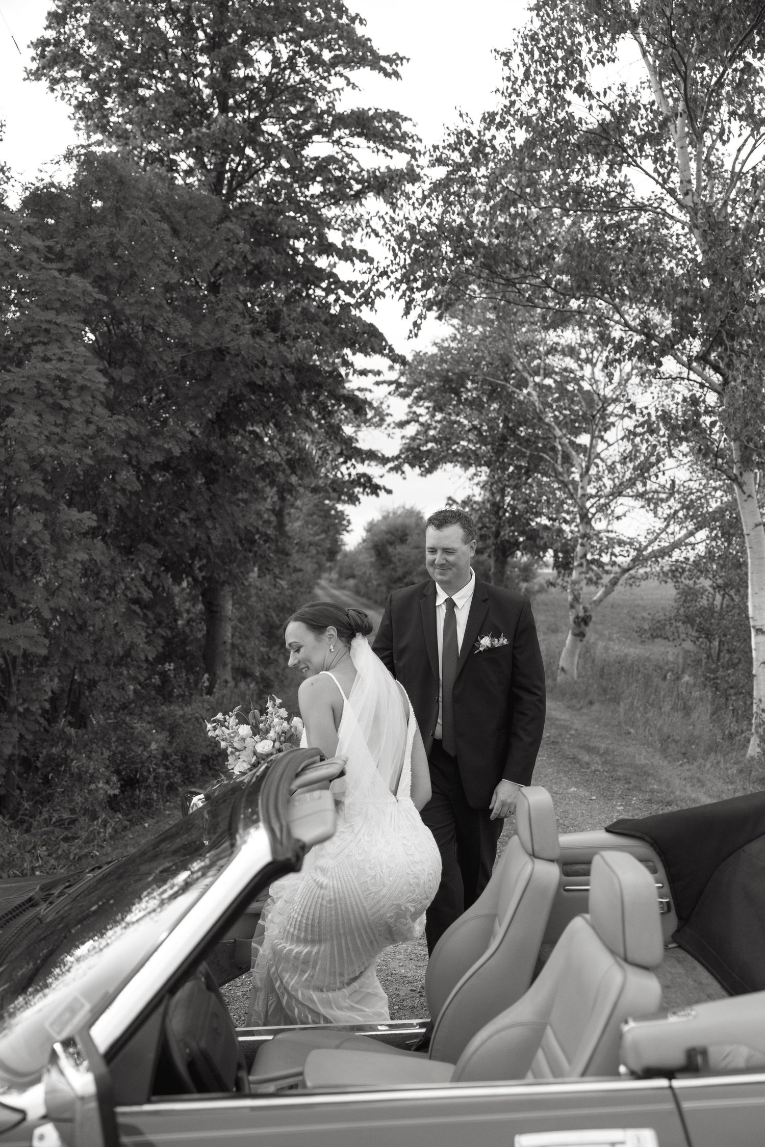 Black and white photo of couple getting into vintage convertible after wedding ceremony.