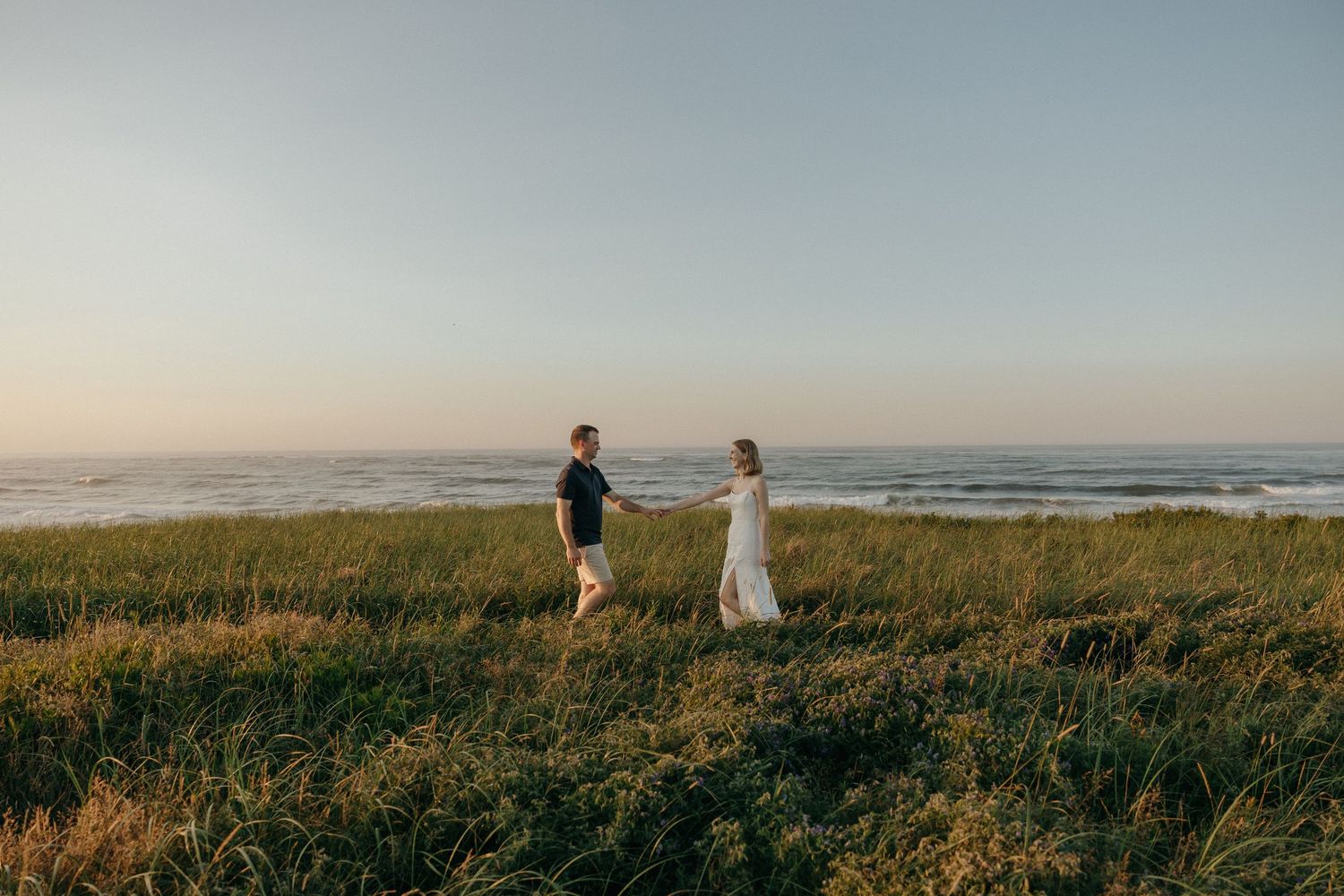 A couple stands together on a grassy coastal cliff overlooking the ocean at sunset.