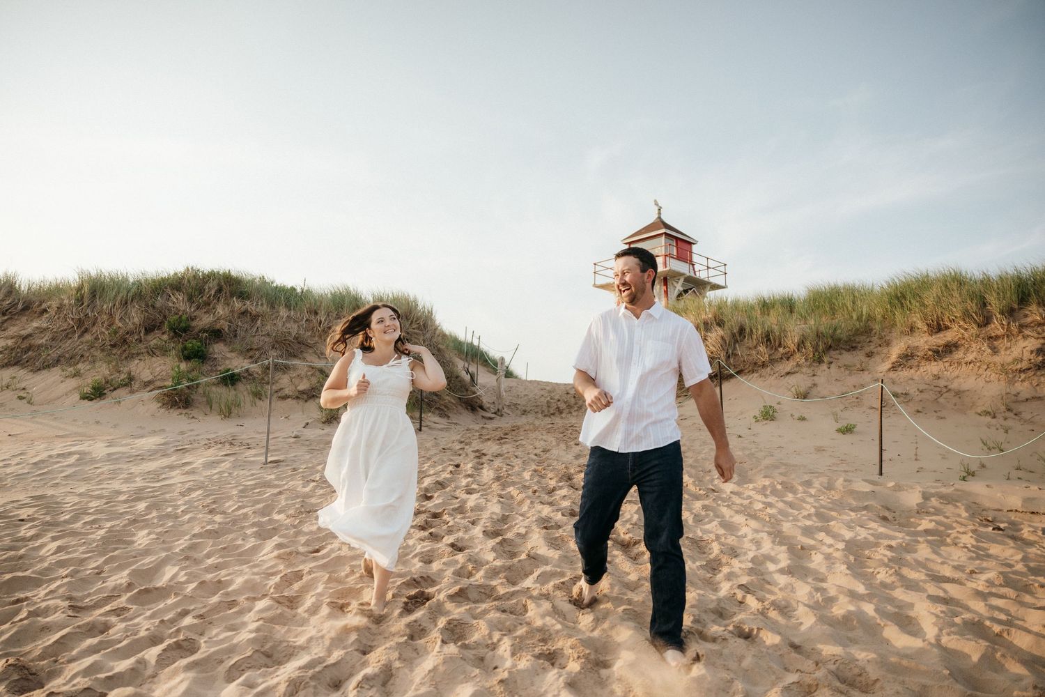 Couple walking hand in hand on sandy beach path near a lighthouse.