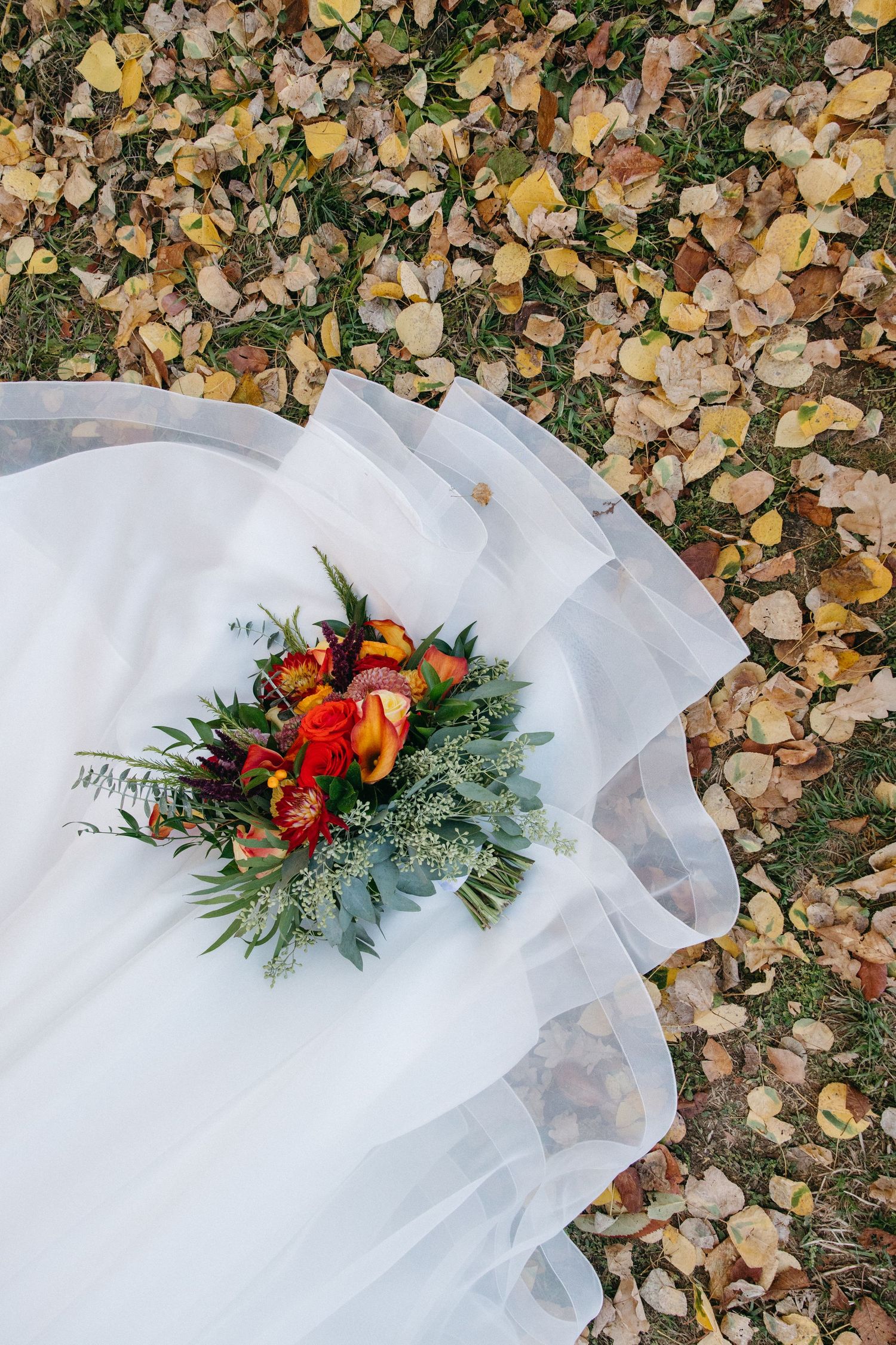 Wedding dress train spread over fallen autumn leaves with vibrant floral arrangement.