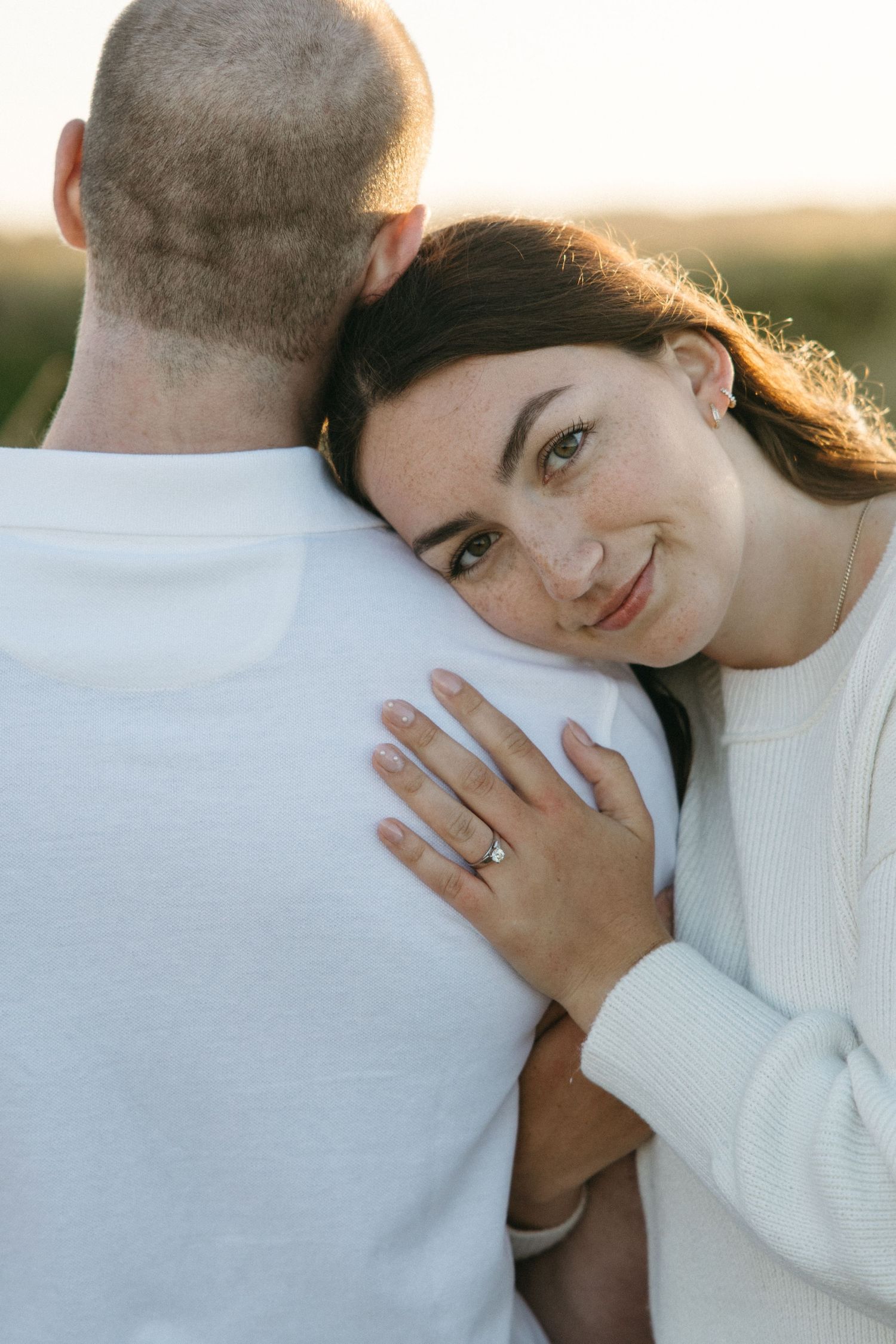 Close-up of engagement ring shown while couple embraces in soft lighting.