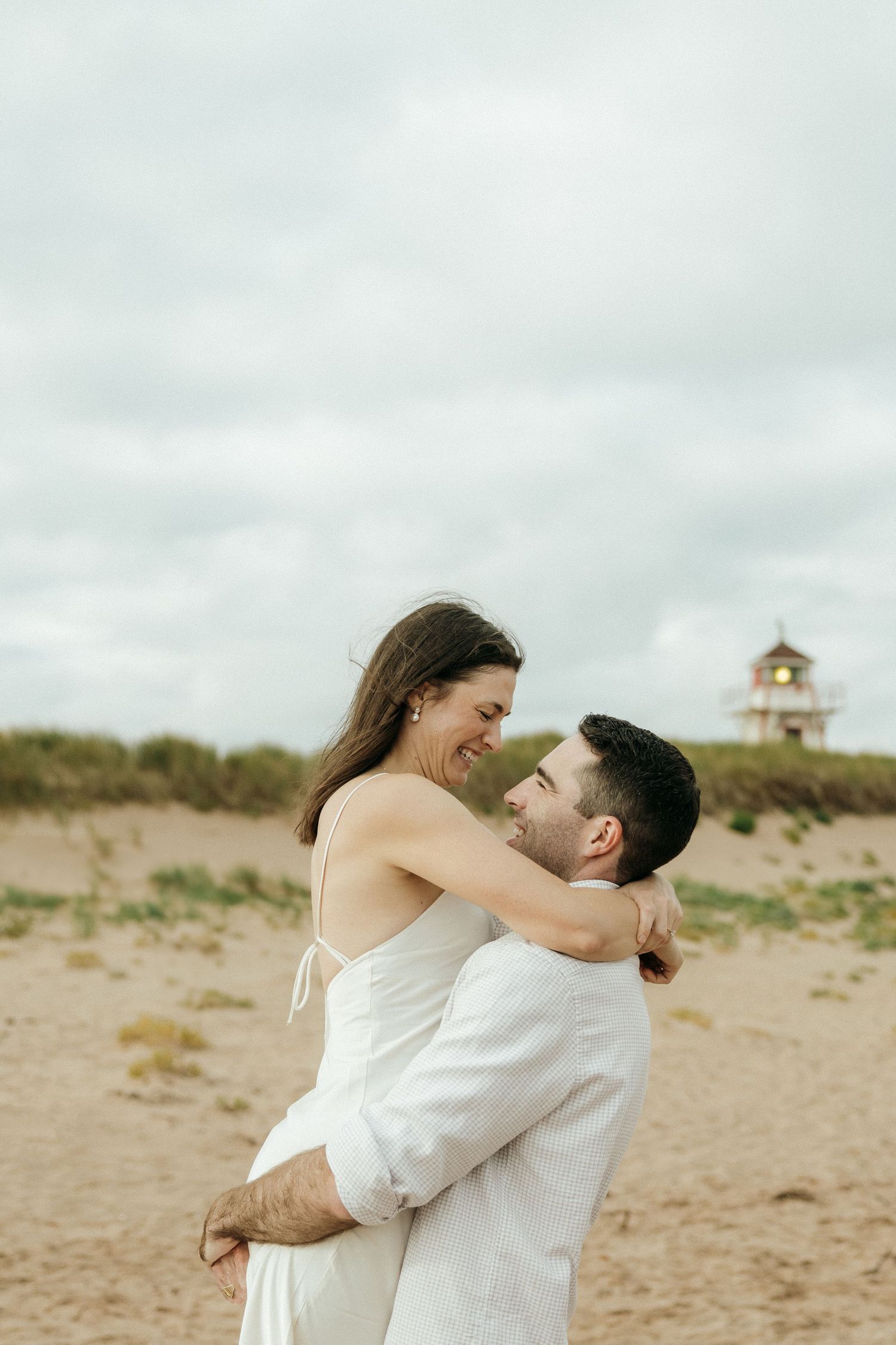 A couple embraces on a beach with a lighthouse and dunes visible in the background.