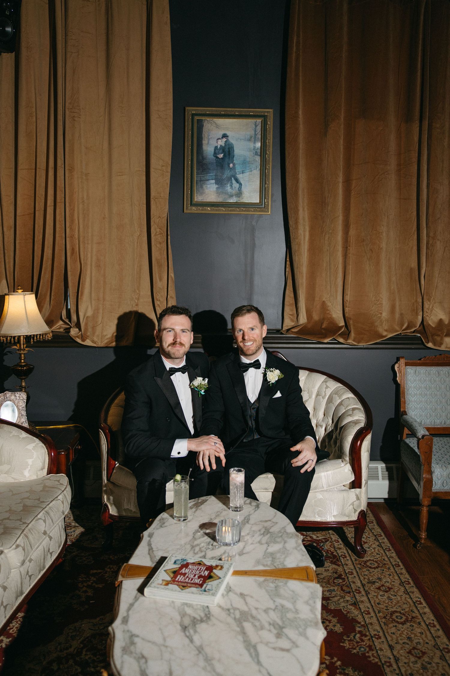 Wedding party members relaxing on vintage furniture in a room with brown curtains and classic decor.