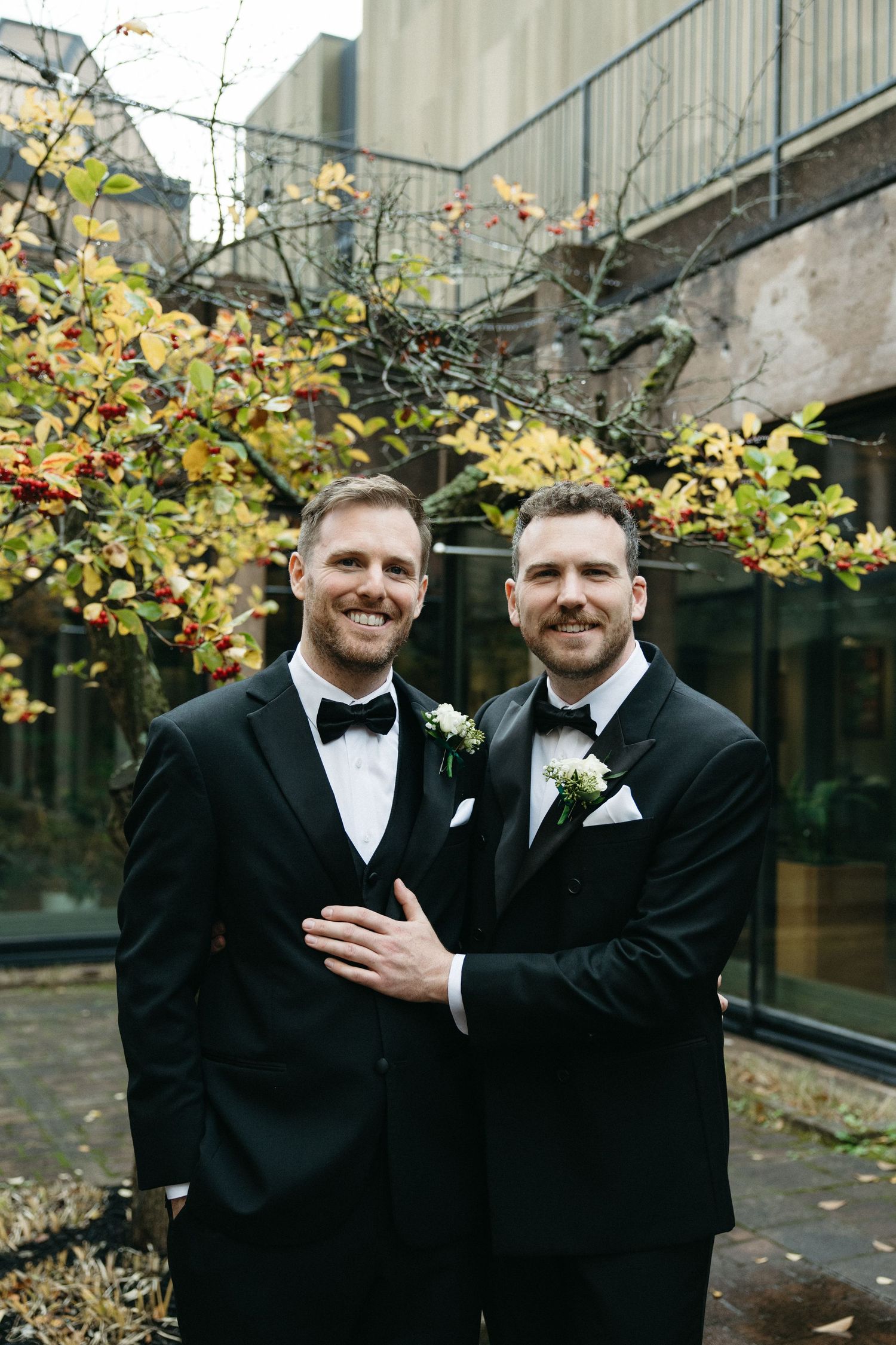 Two people in black tuxedos share joyful moments among autumn foliage.