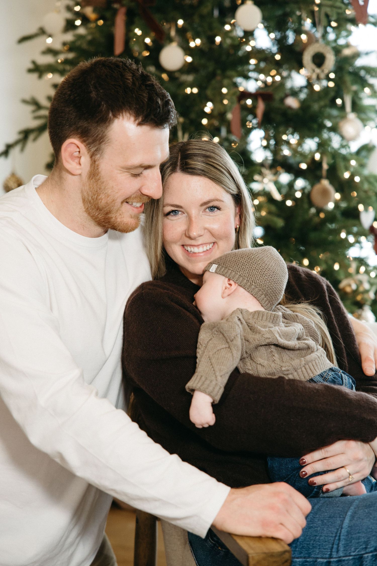 A couple shares tender moments in front of a decorated Christmas tree, wearing white and black sweaters.
