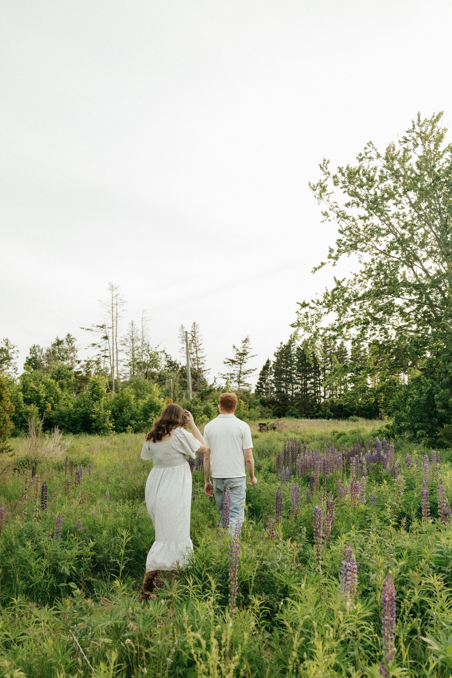 A couple strolls through a lush meadow filled with purple wildflowers and tall grass on a summer evening.