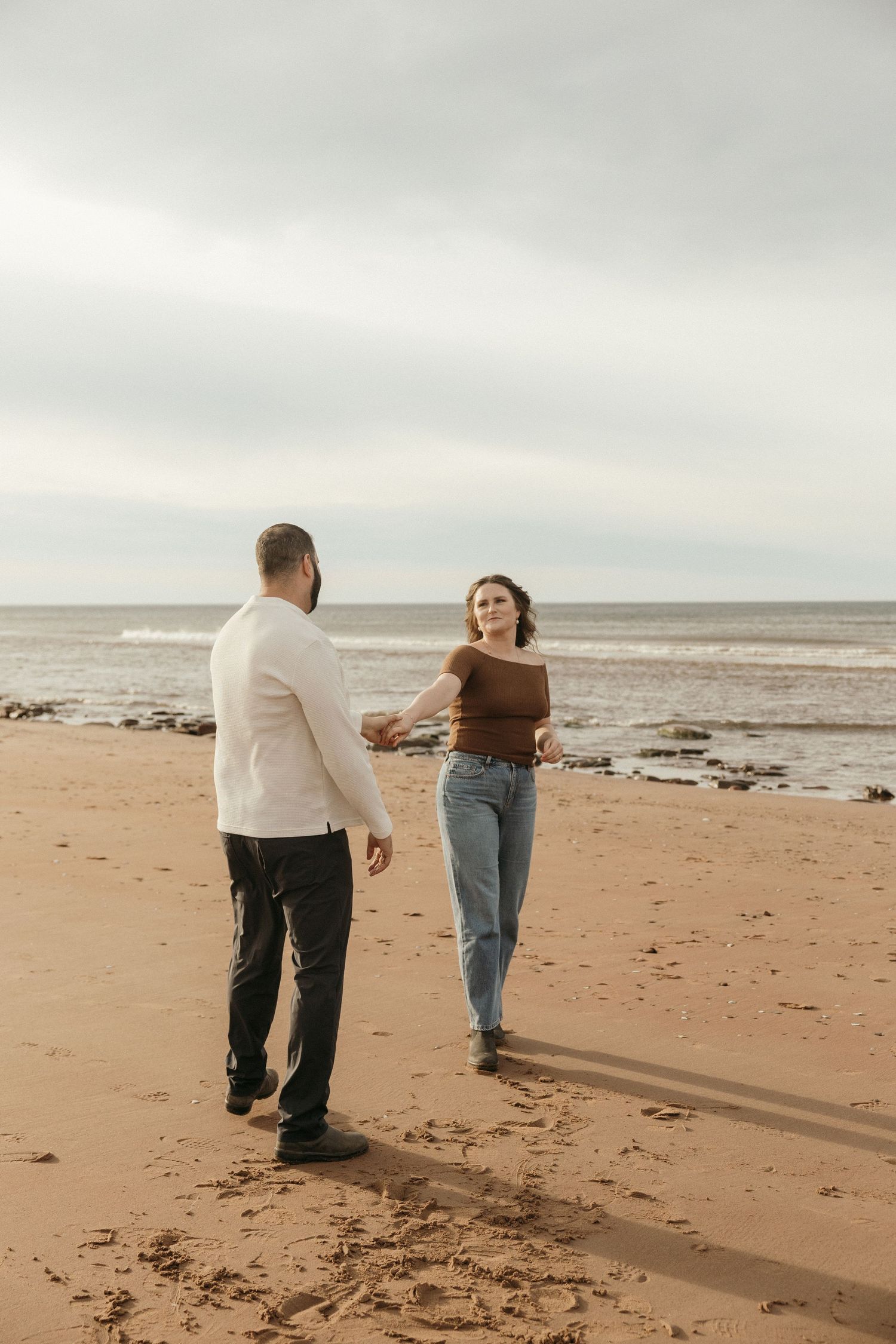 A couple dances playfully together on a sandy beach with the ocean and cloudy sky in the background.