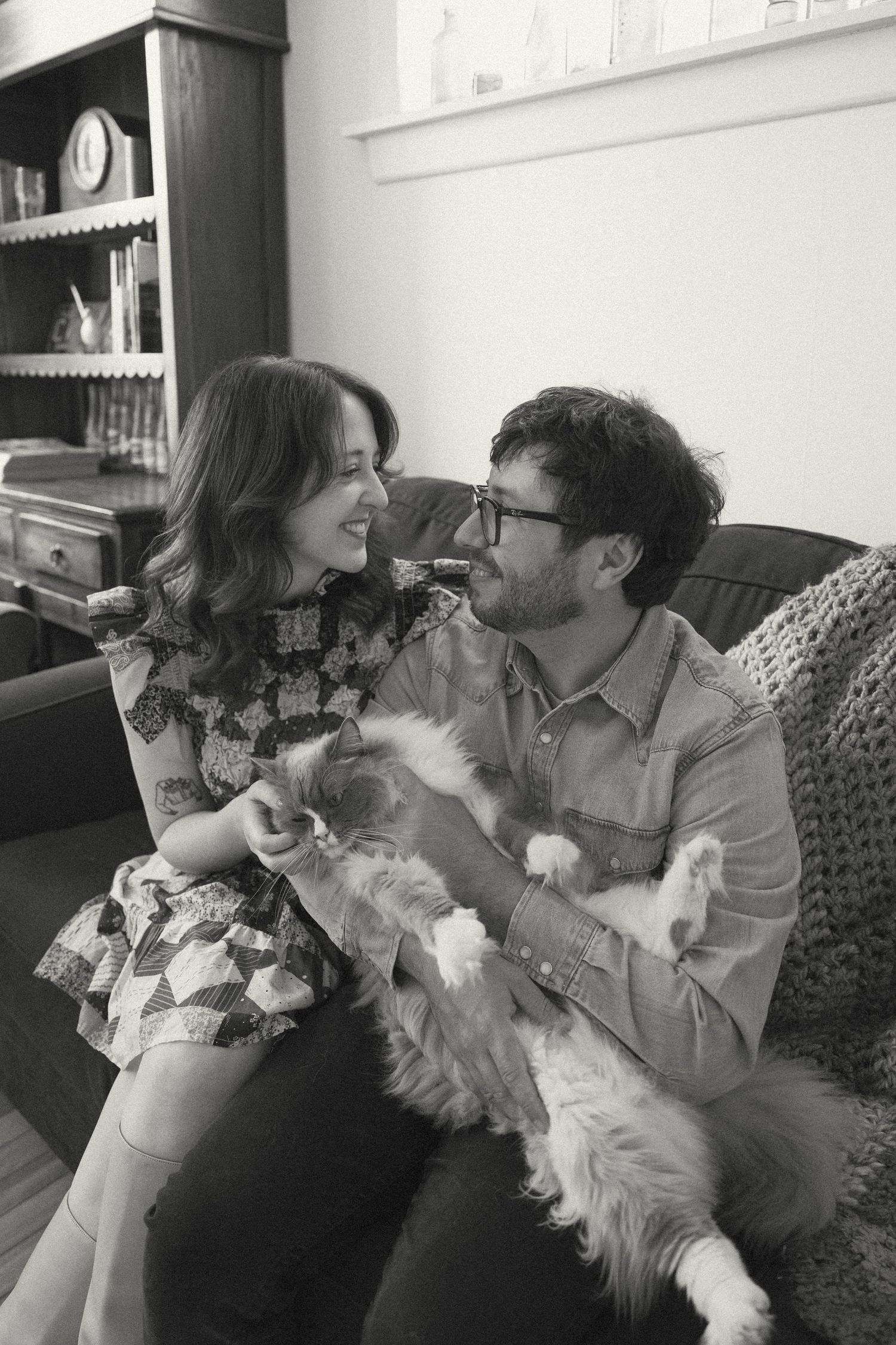 Black and white photo of a smiling couple sitting on a couch together holding a fluffy white cat.