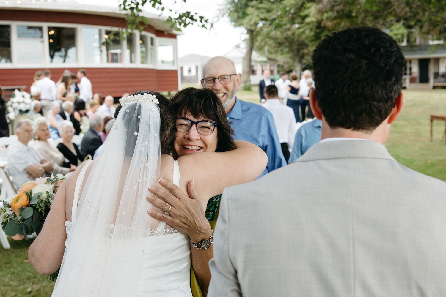 Joyful woman hugging bride in white veil at outdoor wedding reception with guests seated in background.