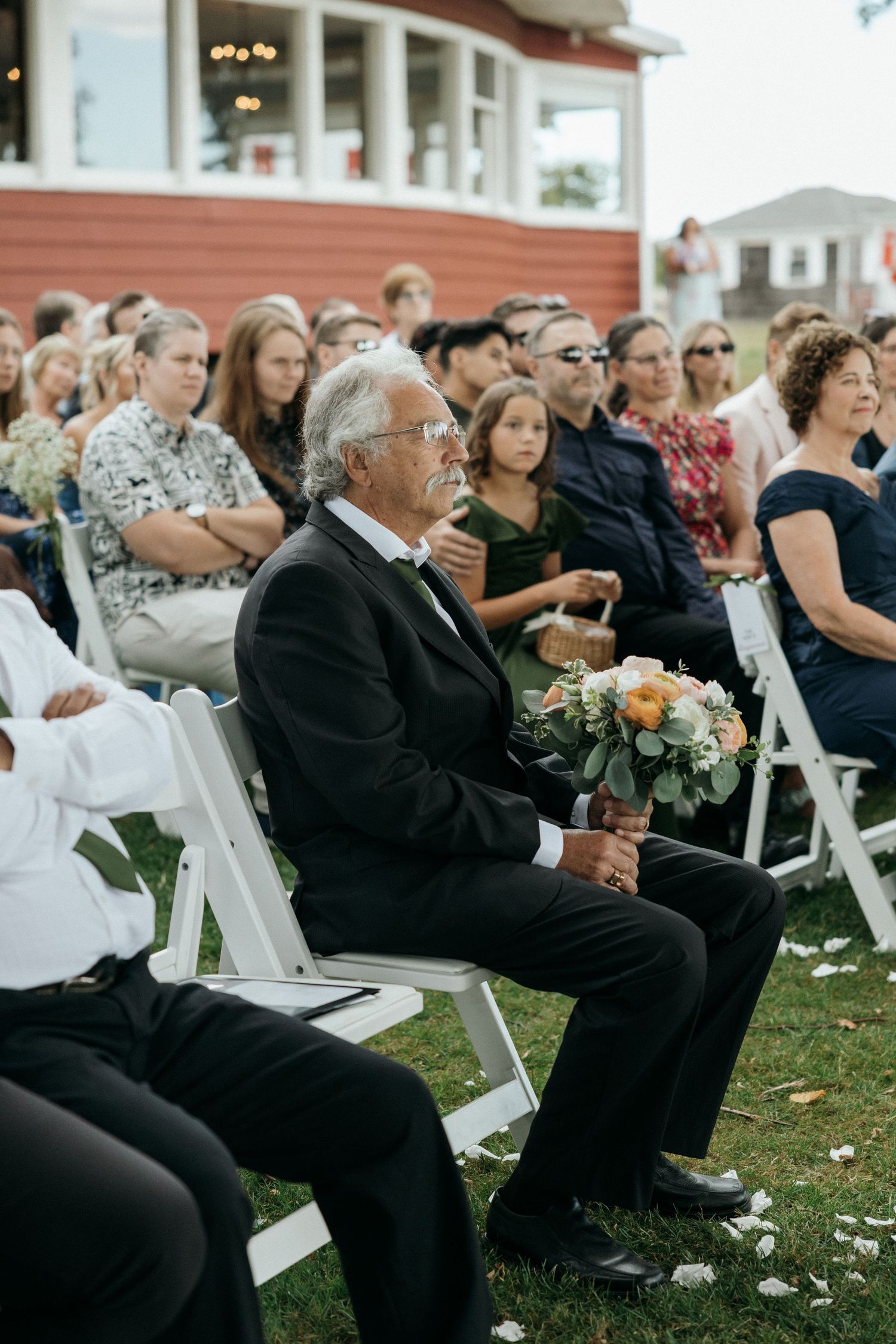Elderly man in black suit holds bridal bouquet while seated at outdoor wedding ceremony on white chairs.