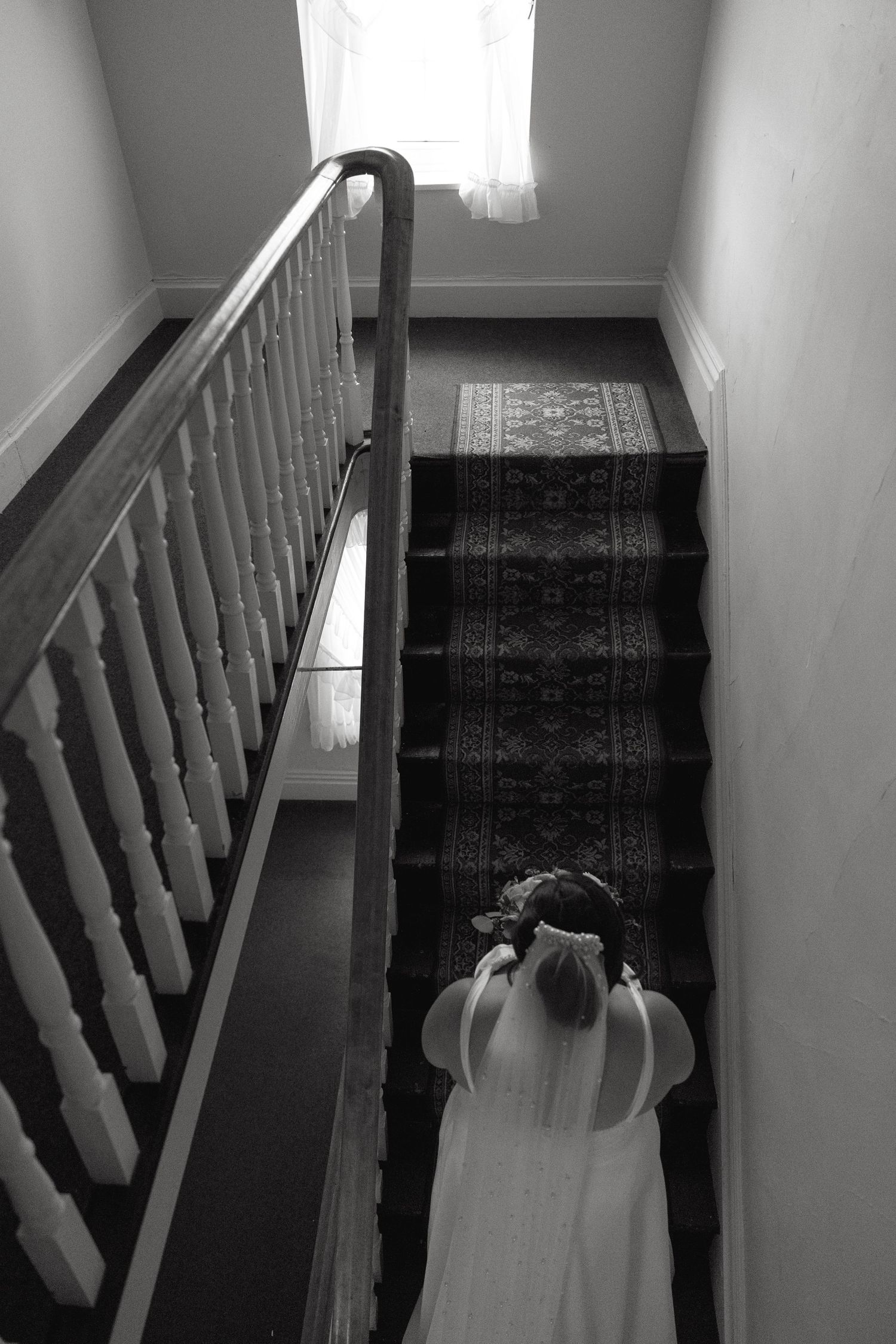 Black and white photo of a bride in a white gown and veil descending a classic wooden staircase.