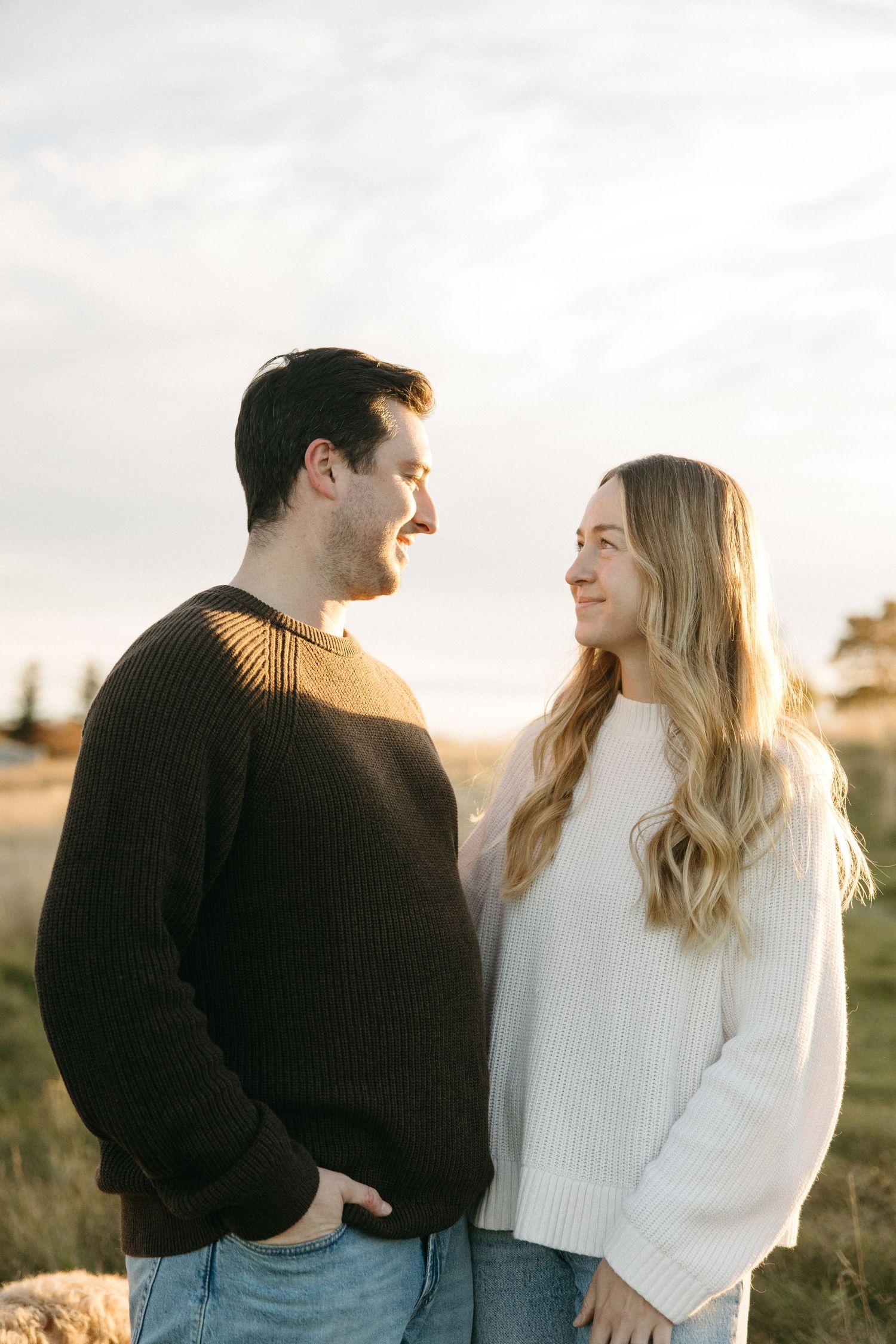 Smiling couple in cozy knit sweaters gazing at each other in a grassy field at golden hour sunset.