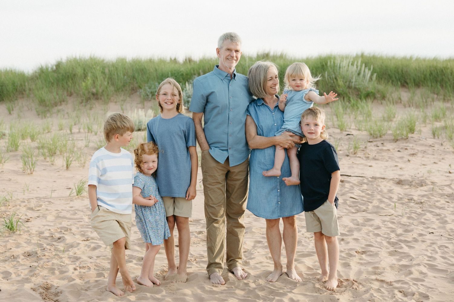 A multigenerational family poses together smiling on a sandy beach with green dunes.