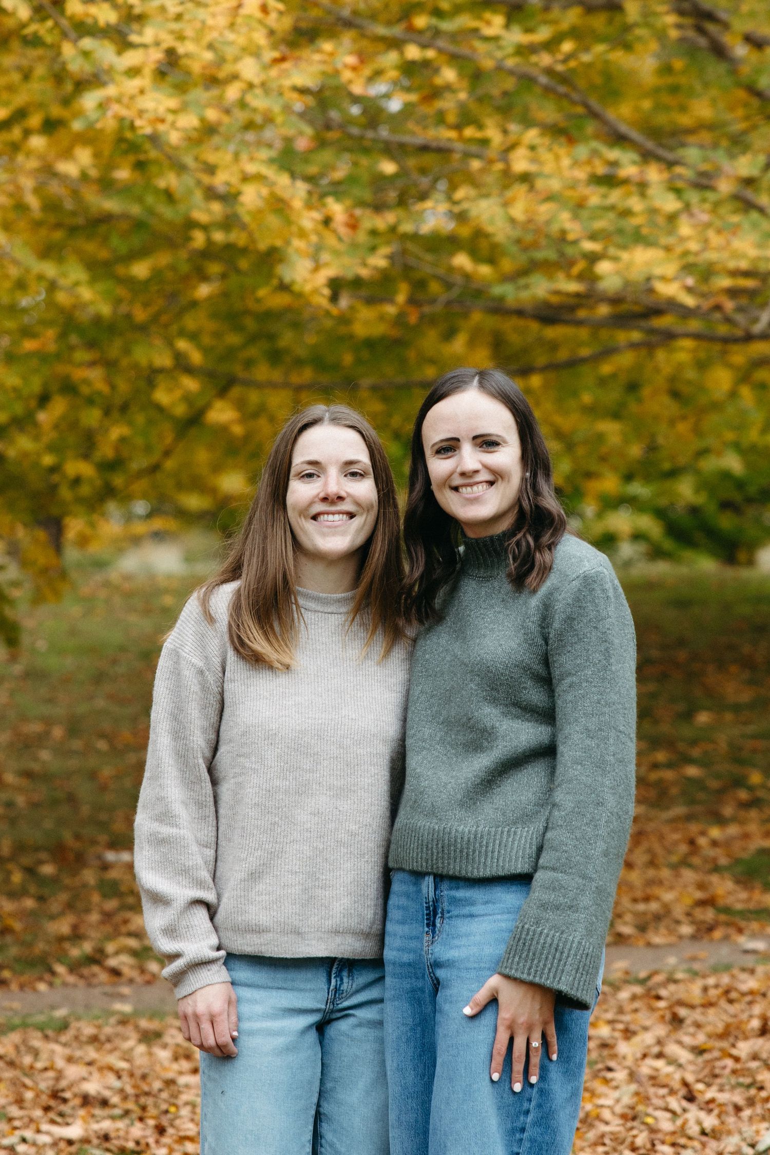 Two smiling women in cozy knit sweaters and jeans pose together under golden autumn trees on a leaf-covered path.