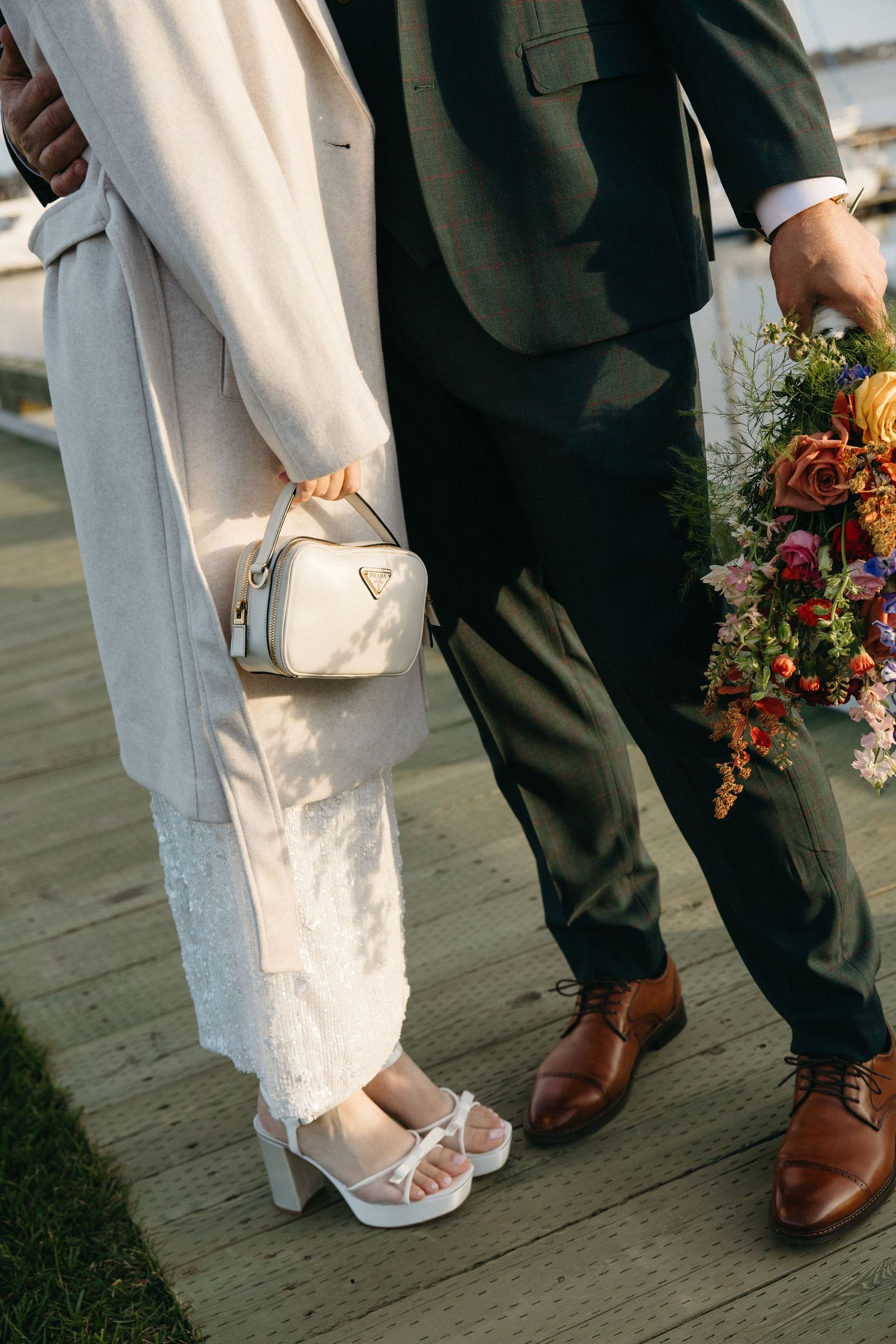 Bride in white lace dress, beige coat and platform heels stands beside a groom in green suit and brown shoes holding a colorful bouquet.