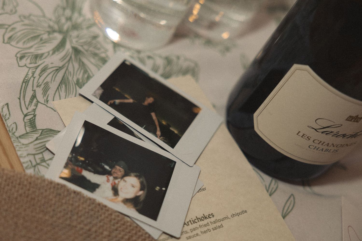 Two instant photos rest on a printed restaurant menu beside a bottle of Champagne with a dark label and a glass on the table.