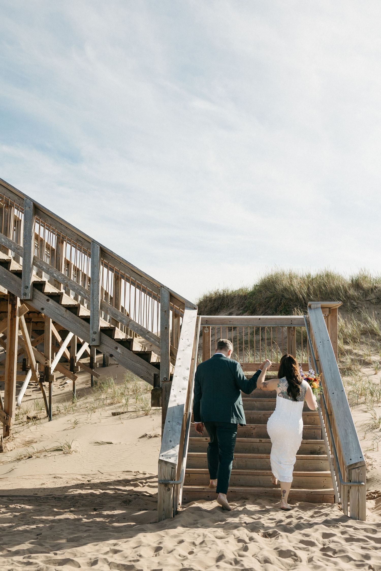 Bride in a white dress and groom in a dark suit walk up wooden beach stairs through sand dunes under a blue sky.