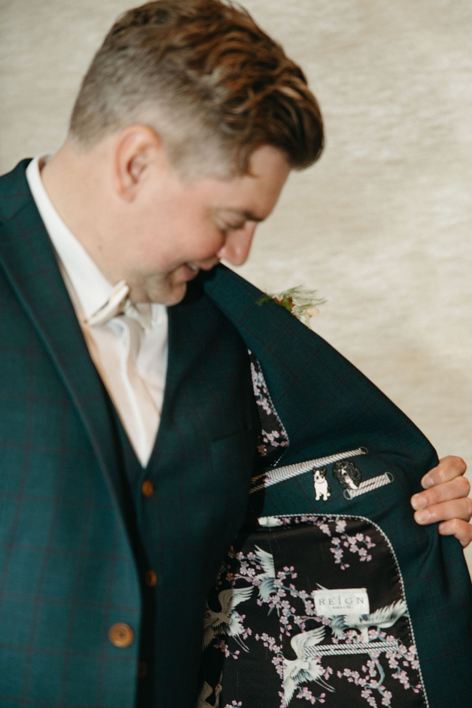 Smiling groom in a teal plaid suit and waistcoat opens his jacket to show a floral bird-print lining with a custom label.
