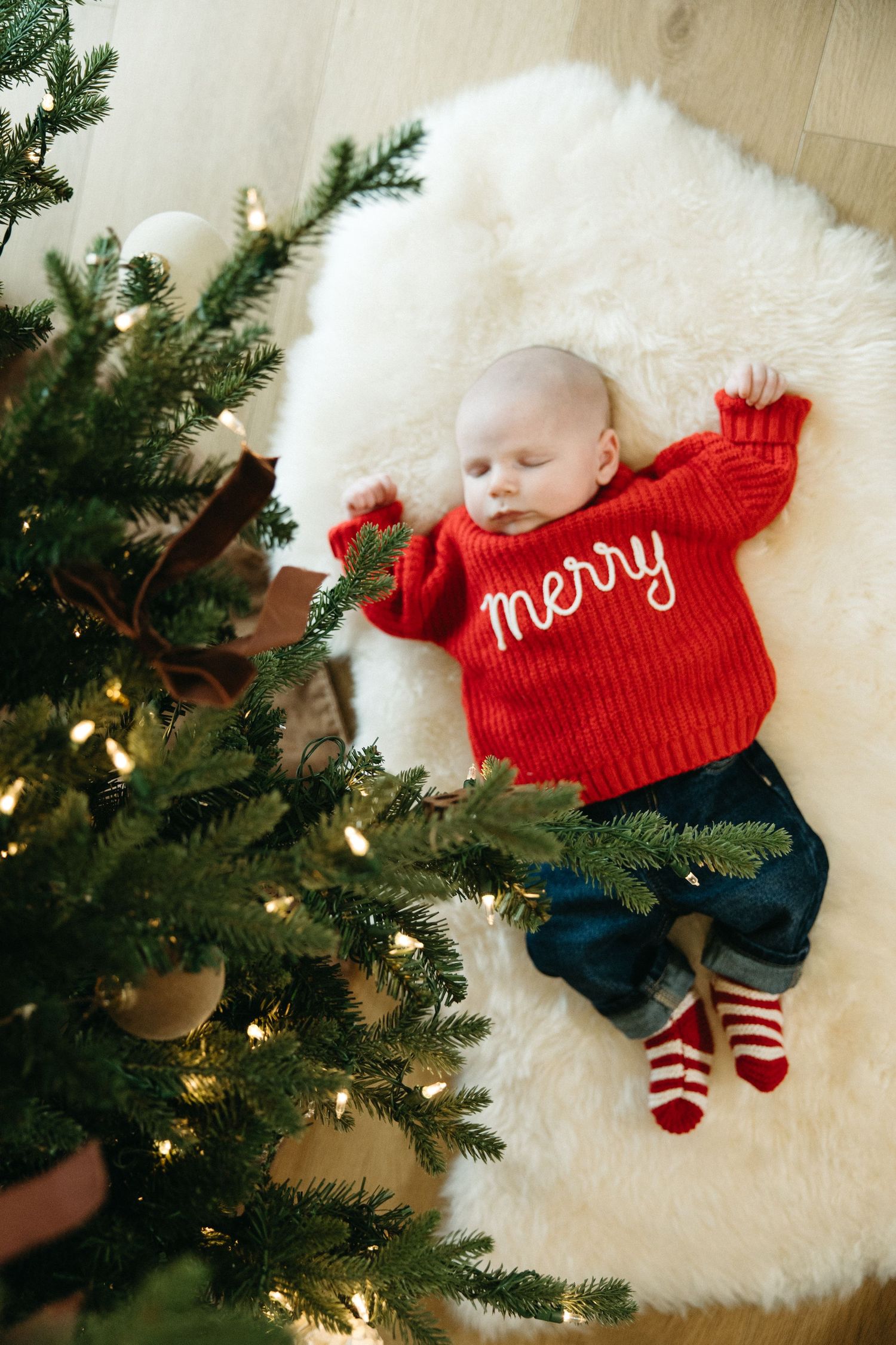 A sleeping baby in a red 'Merry' sweater and striped socks rests under a lit Christmas tree.