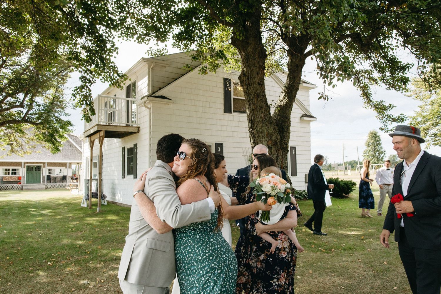 Wedding guests hugging and socializing outdoors near a white farmhouse surrounded by trees on a sunny day.